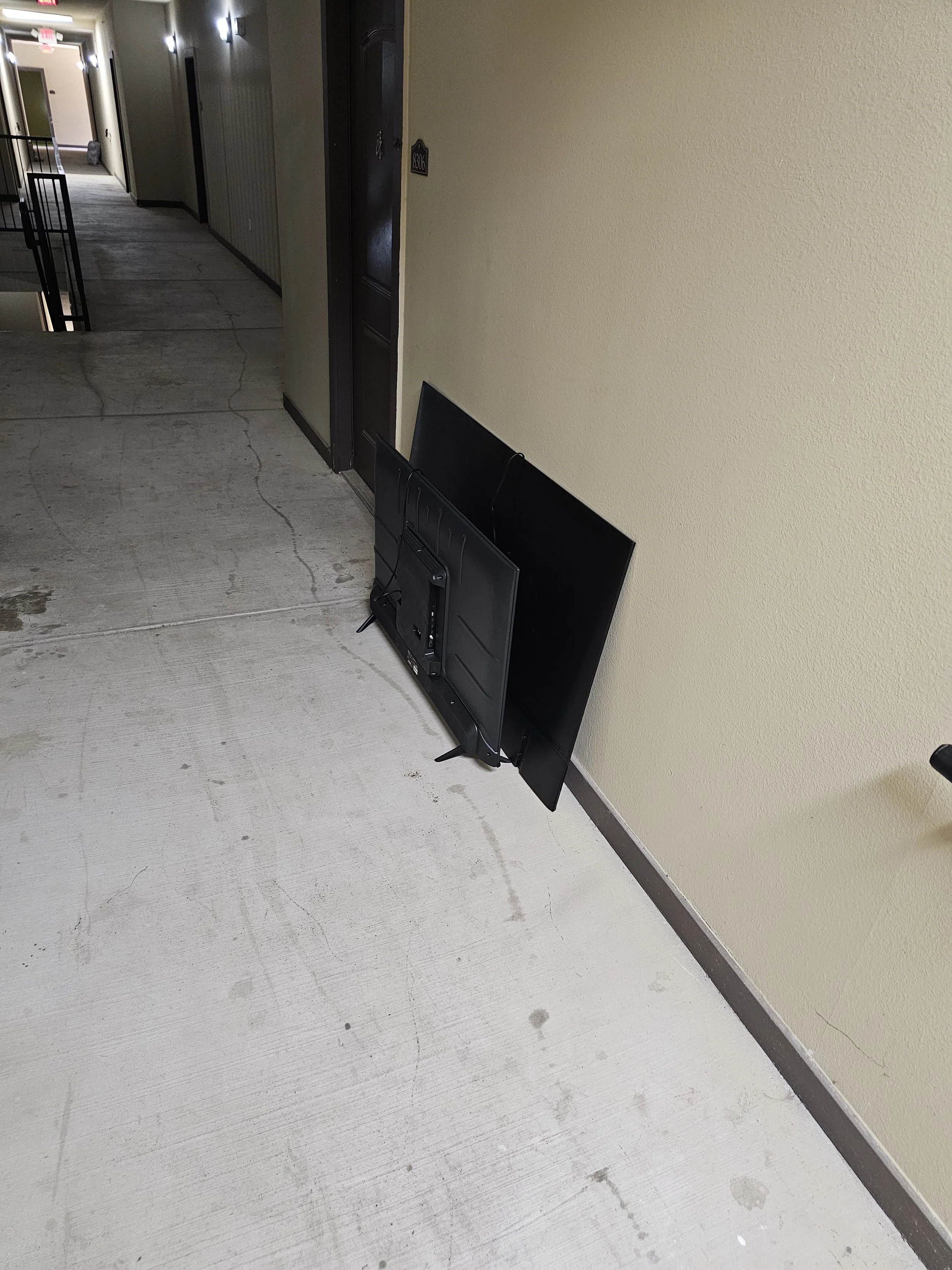 An outdoor apartment hallway with a wall-mounted television and a large flat-screen monitor leaning against the wall.