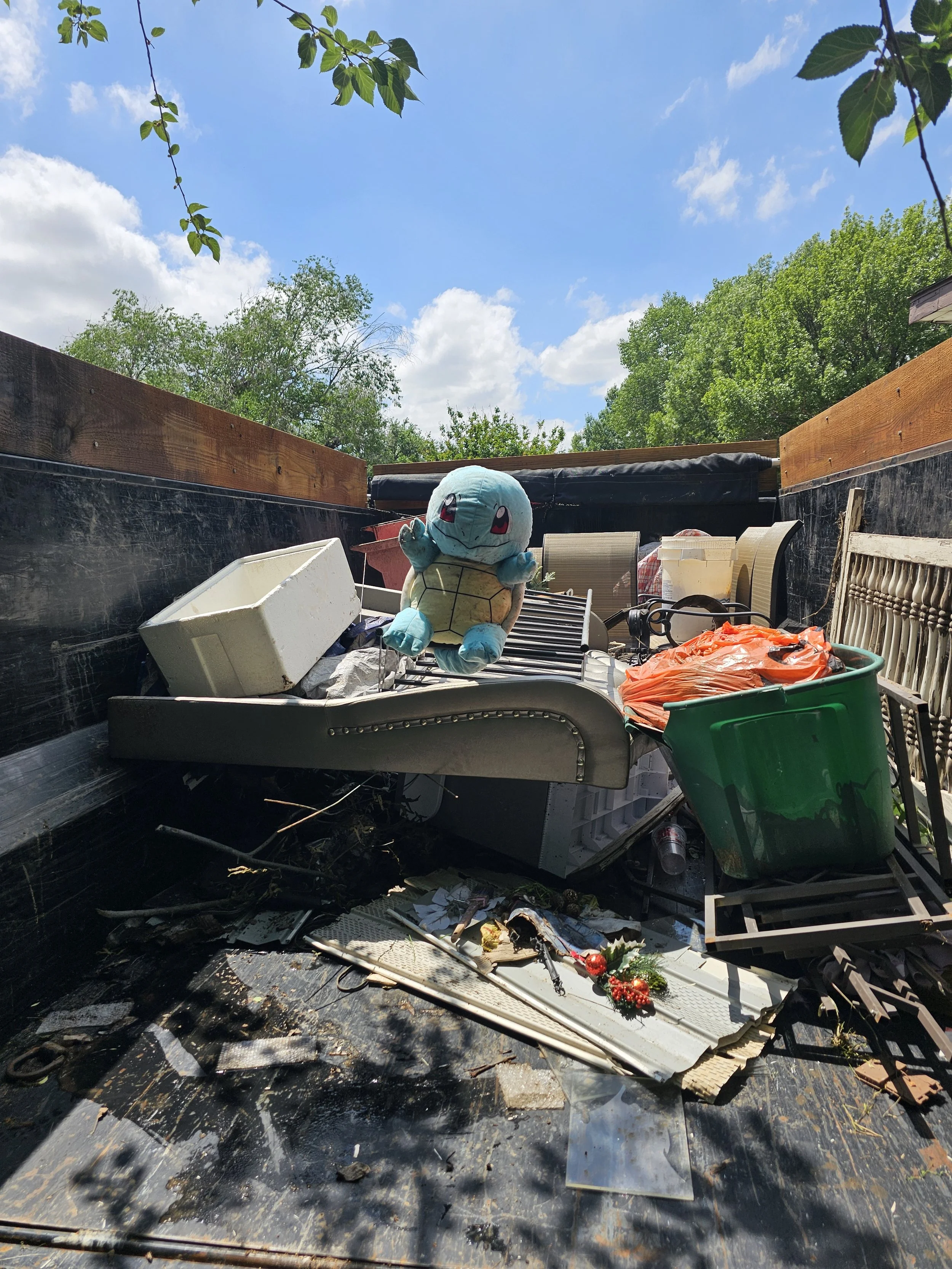 Pickup truck bed filled with discarded household items including a Snorlax plush toy, a green storage bin, a white rectangular object, a beige lamp, and miscellaneous debris under a blue sky with scattered clouds and green trees.