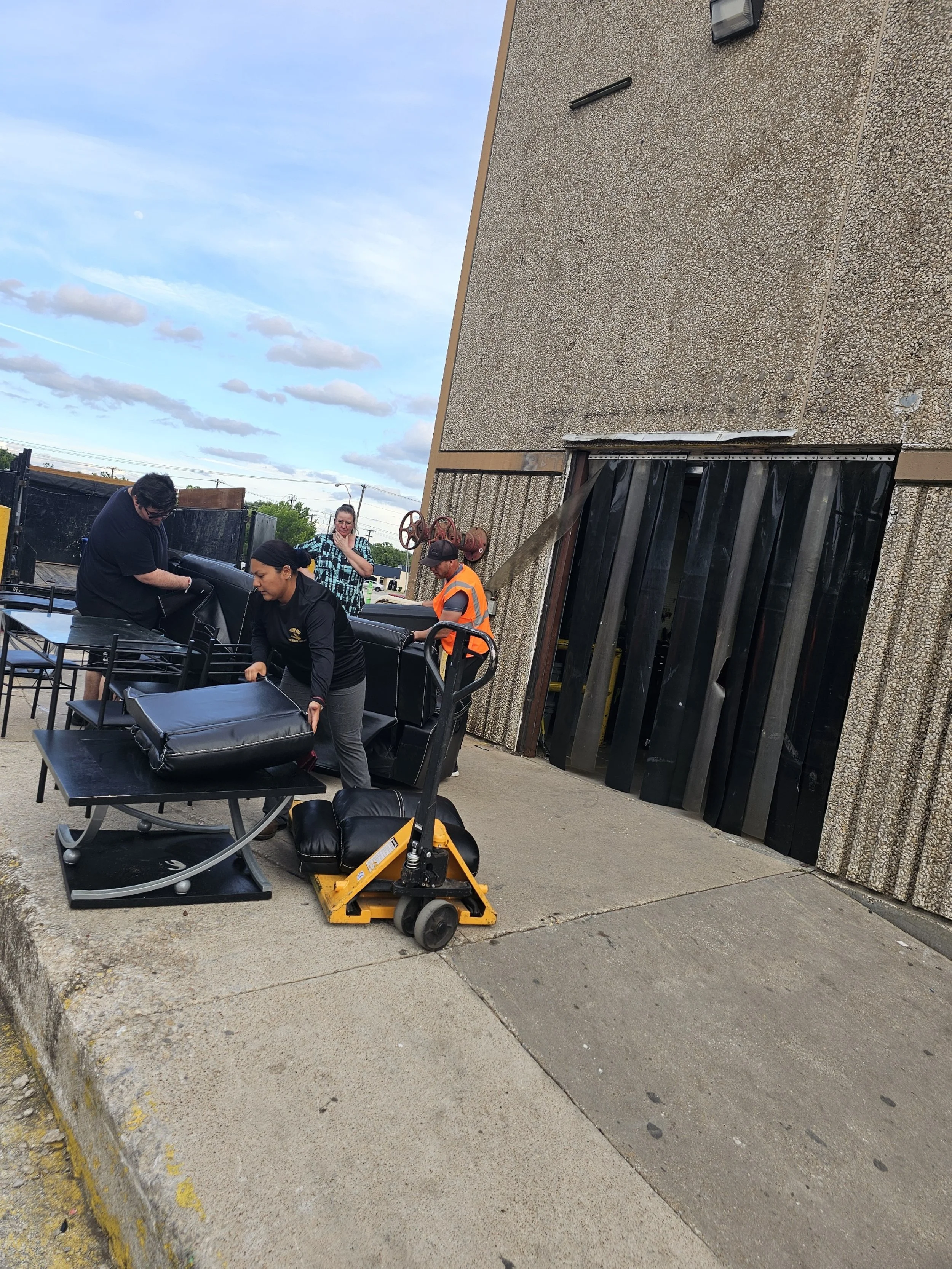 Workers unload black furniture, possibly sofas, outside a building with a textured exterior wall. One worker in an orange safety vest operates a pallet jack, while others assist with the furniture. The sky is partly cloudy.