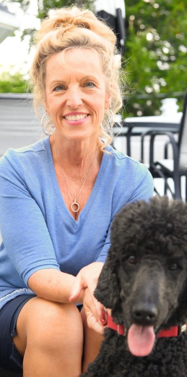 A smiling woman sitting outdoors with a black poodle puppy, greenery in the background.