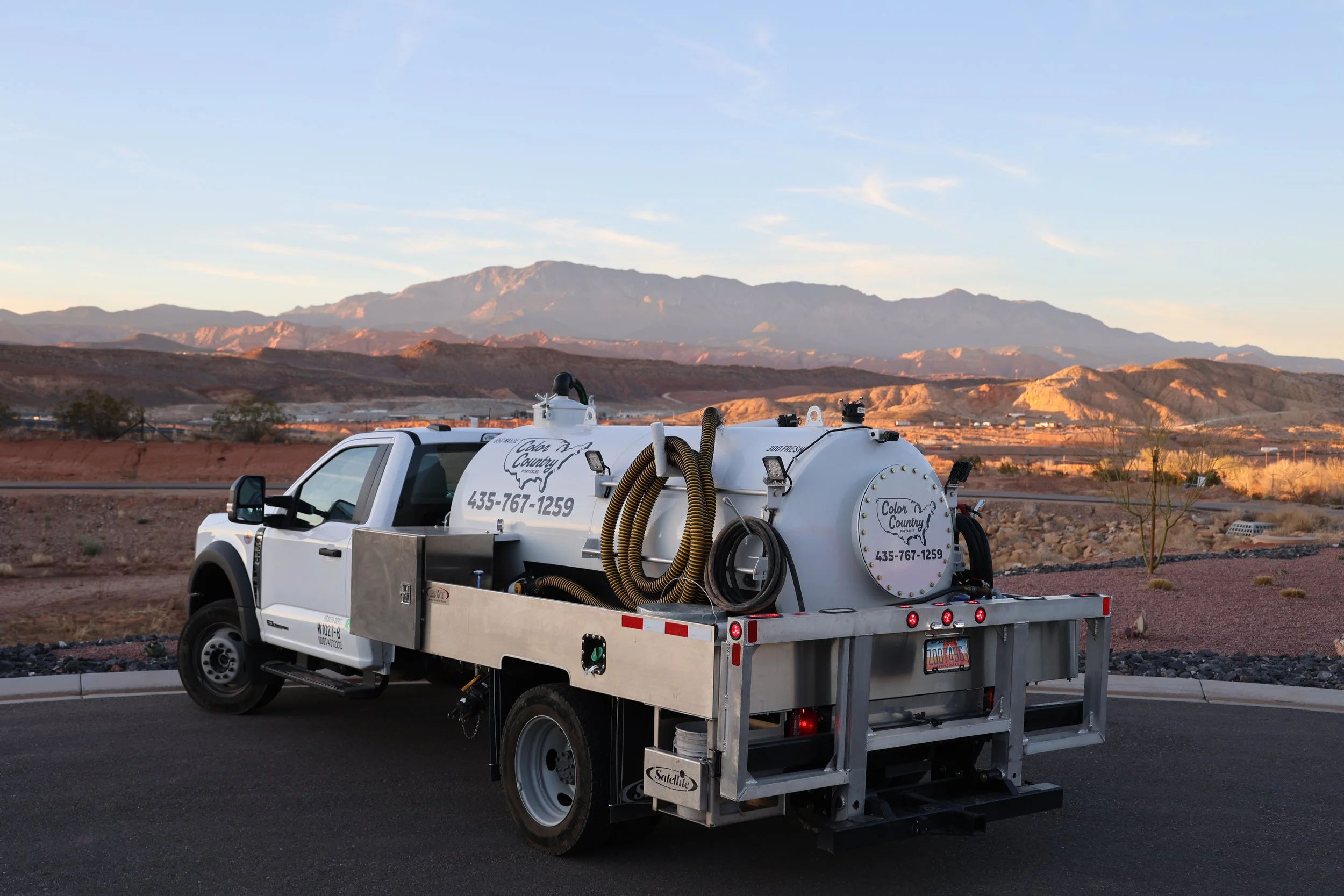 A white serviced truck with equipment on its bed, parked on a desert highway with mountains in the background during sunset.