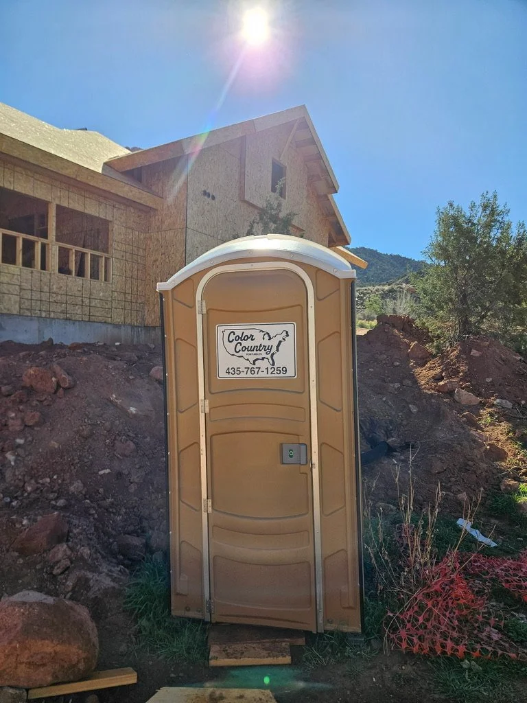 A portable toilet with a sign that reads 'Color Country Portables' and a phone number, set outdoors in front of a house under construction with a clear blue sky and the sun shining.