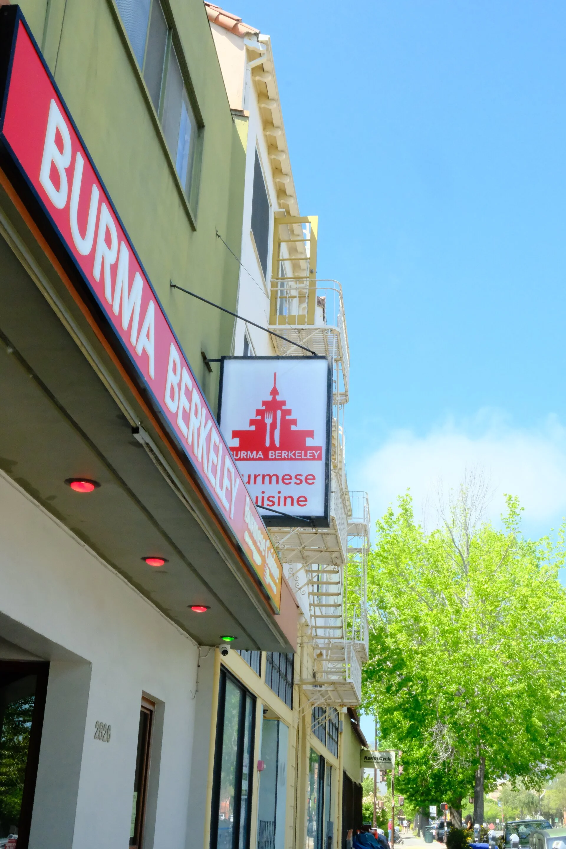 Street view of a building with a sign for Burmese cuisine, featuring a red pagoda-like logo and text 'Burma Berkeley' on a white background. The building has a green facade, and there are metal fire escapes on an adjacent building. Bright blue sky and green trees are visible in the background.