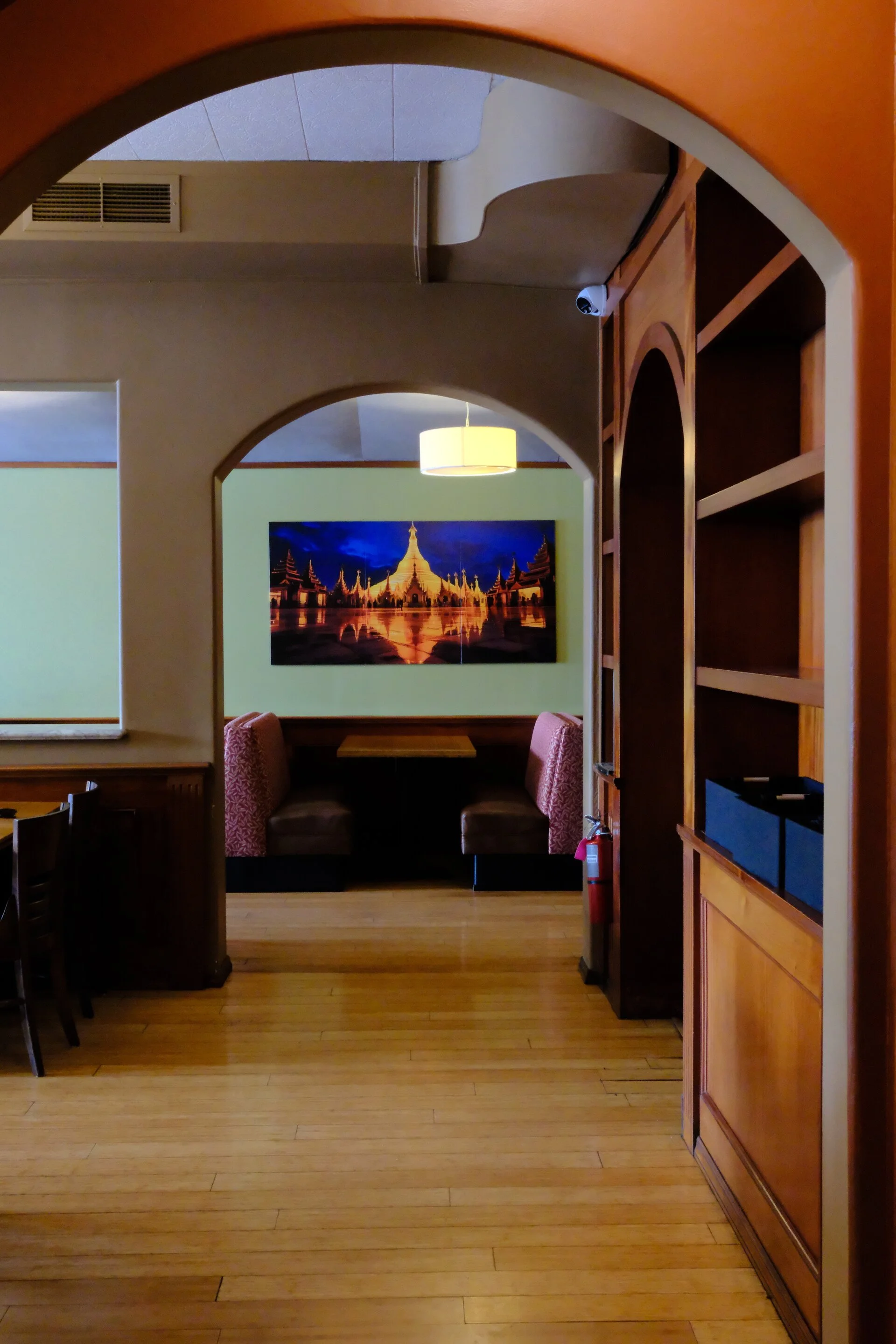 Interior view of a restaurant corridor with wooden floors, arched doorways, a framed illuminated picture of a temple, and a seating area with pink upholstered booths.