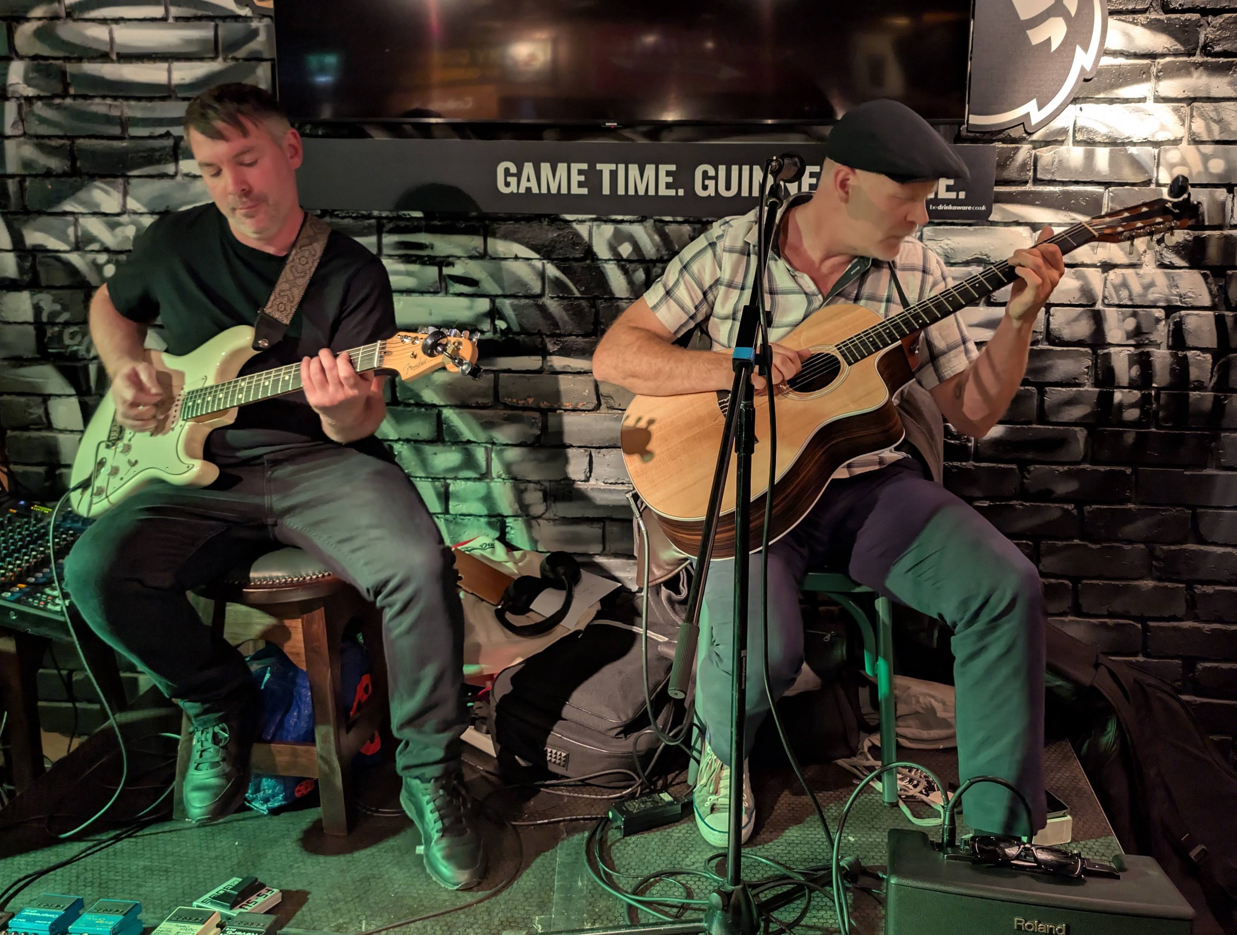 Two men playing guitars on a small stage with a black brick background, one acoustic and one electric guitar, in a dimly lit venue.