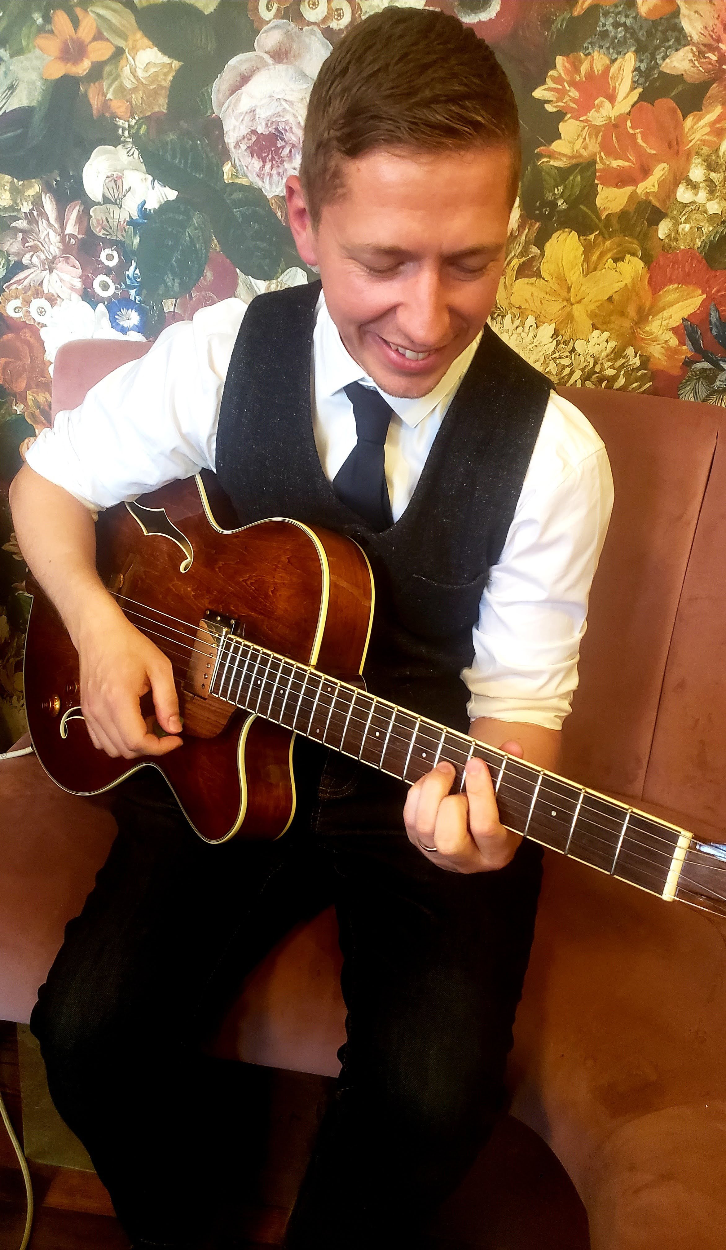 A smiling man in a white shirt, black vest, and black tie playing an acoustic guitar in front of a floral wallpaper background.