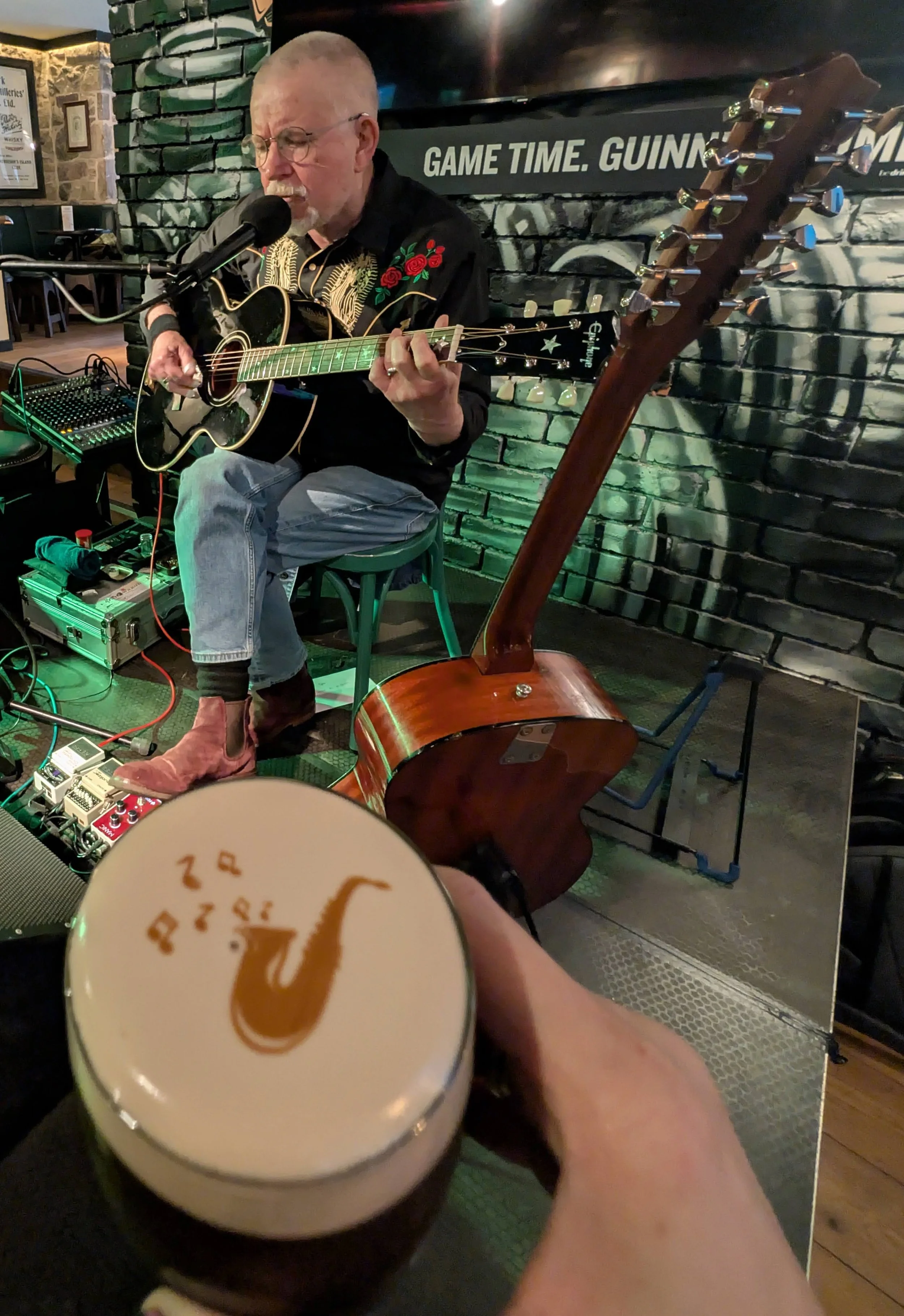 A man playing an acoustic guitar and singing into a microphone in a bar or pub setting, with a brick wall background and a sign that says 'Game Time. Guinness.' in the background. Someone is holding a pint of beer with a saxophone logo on it in the foreground.