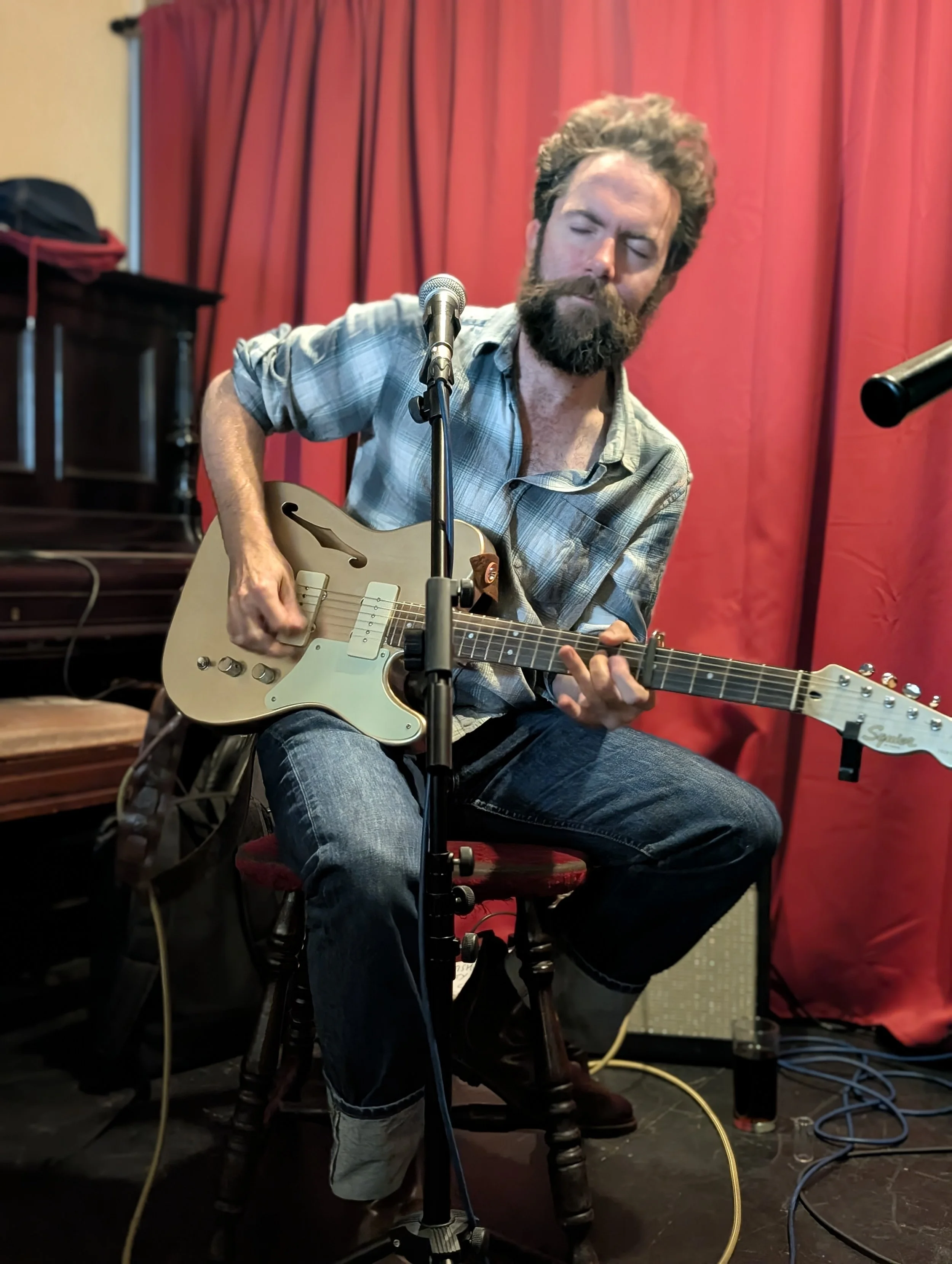 A man with a beard and mustache playing an electric guitar, sitting on a red cushioned stool in a room with red curtains and an amplifier nearby.