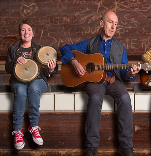 A woman smiling while playing bongos and a man playing an acoustic guitar, sitting on a bench in front of a wooden wall with abstract carvings.