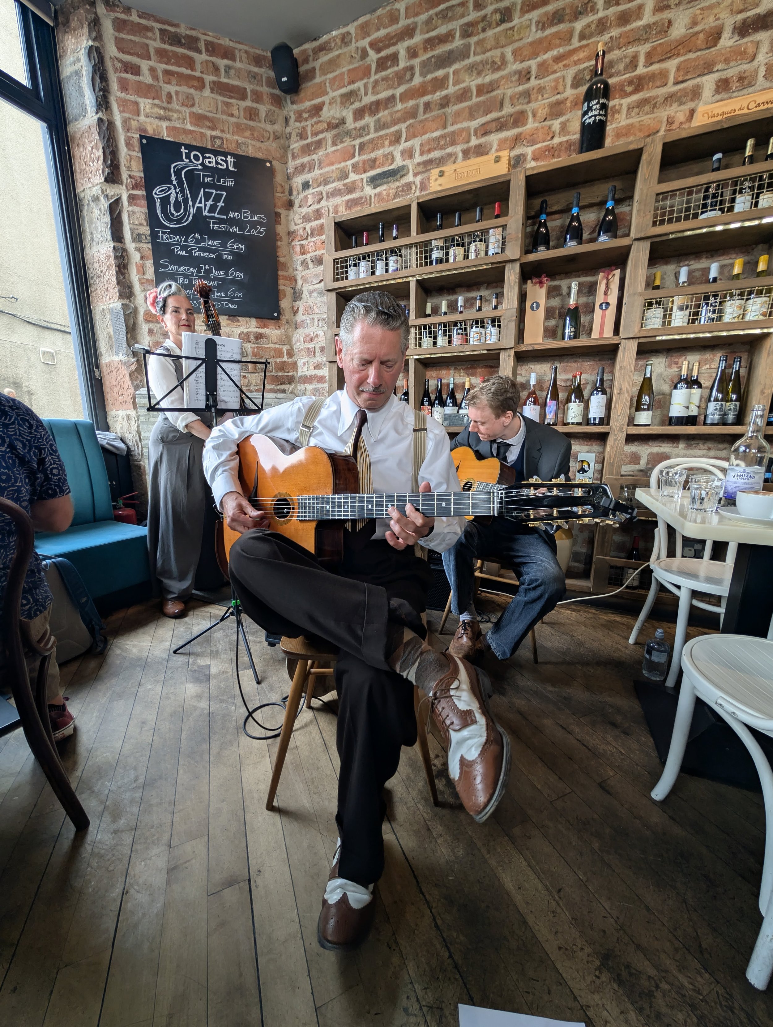 Two men in vintage attire playing acoustic guitars at a cozy café with brick walls, wine bottles on shelves, and a chalkboard advertising jazz and blues festival.