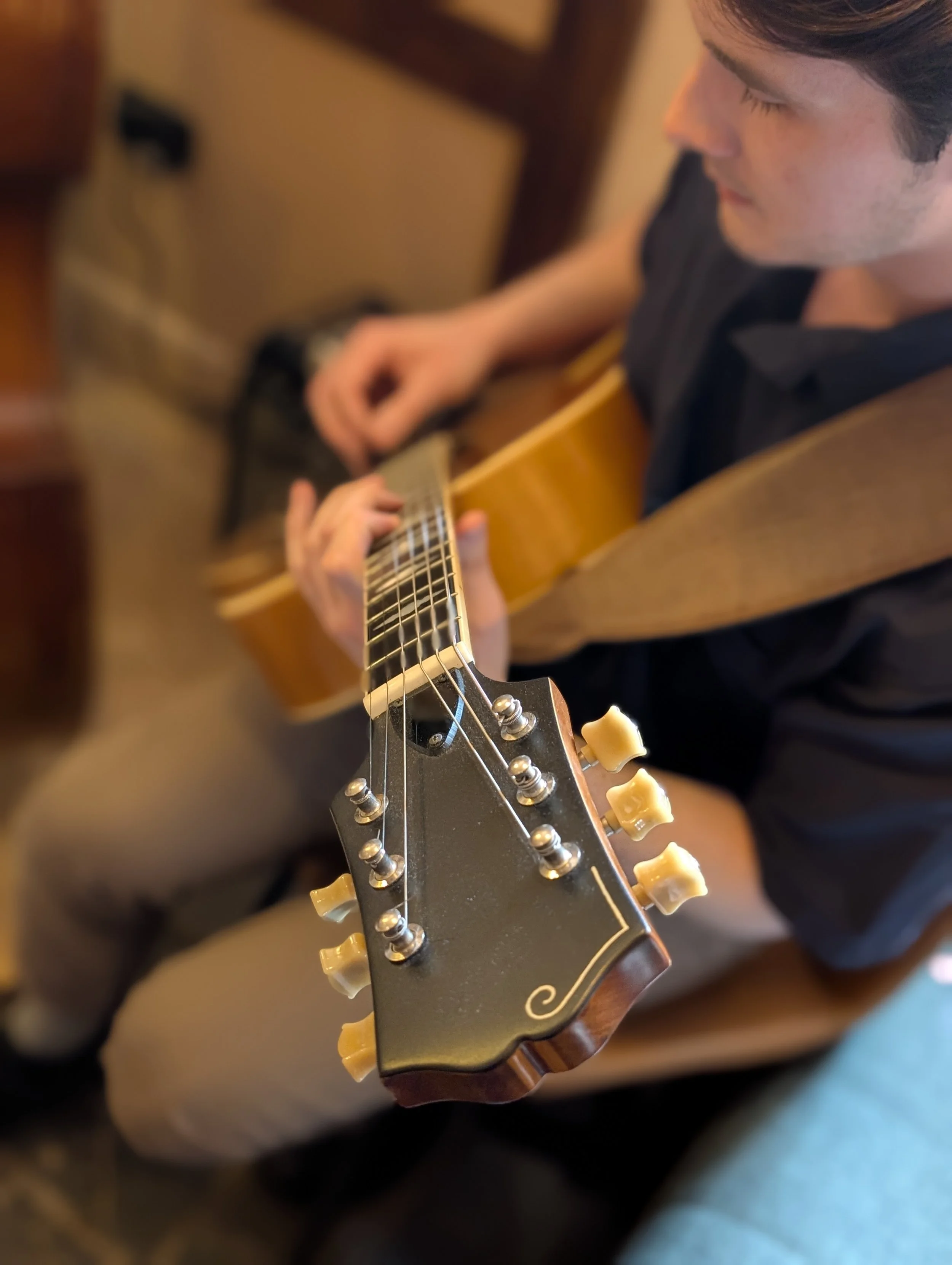 Close-up of a person playing an acoustic guitar, focusing on the guitar's headstock and tuning pegs. The person's face is partially visible, with eyes closed, wearing a dark shirt, sitting in a cozy indoor setting.
