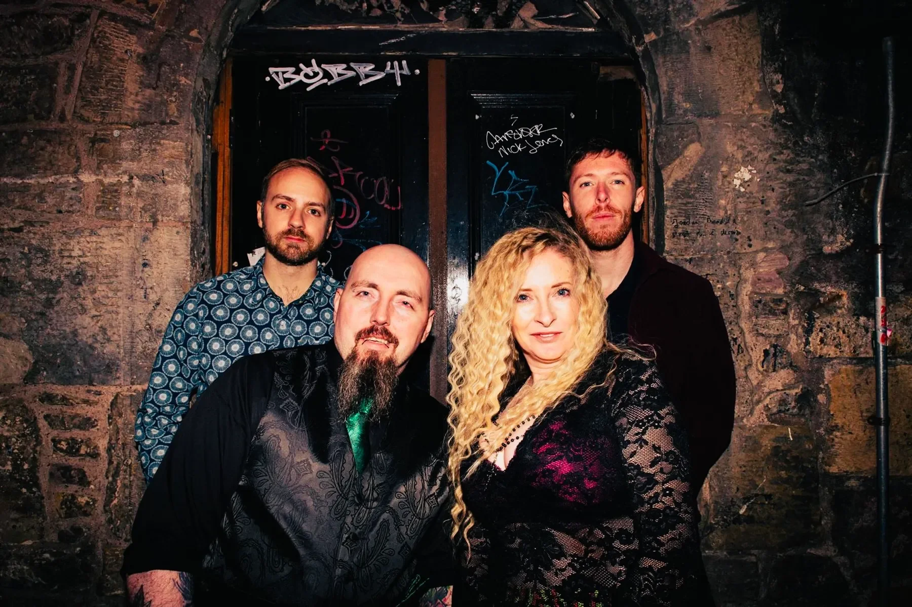 Four people posing in front of a graffiti-covered brick wall. Two men are standing in the back, and a man and woman are in the front. The woman has long curly blonde hair, and the man in front has a beard and mustache. The two men in the back have short hair and beards. The group appears to be at night or in a dimly lit setting.