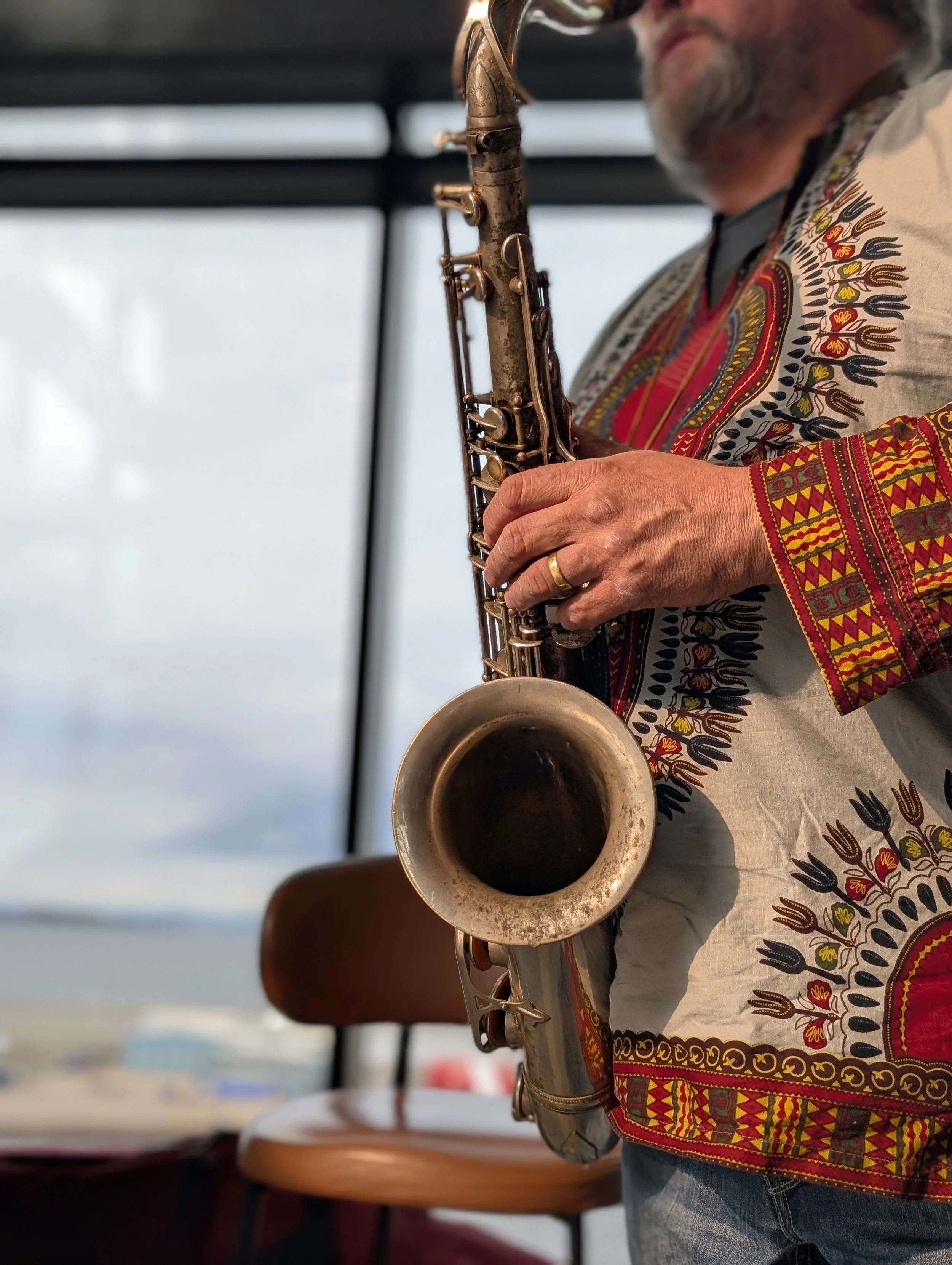 A man playing a saxophone indoors with a large window showing an outdoor scene in the background, wearing a colorful, patterned shirt.