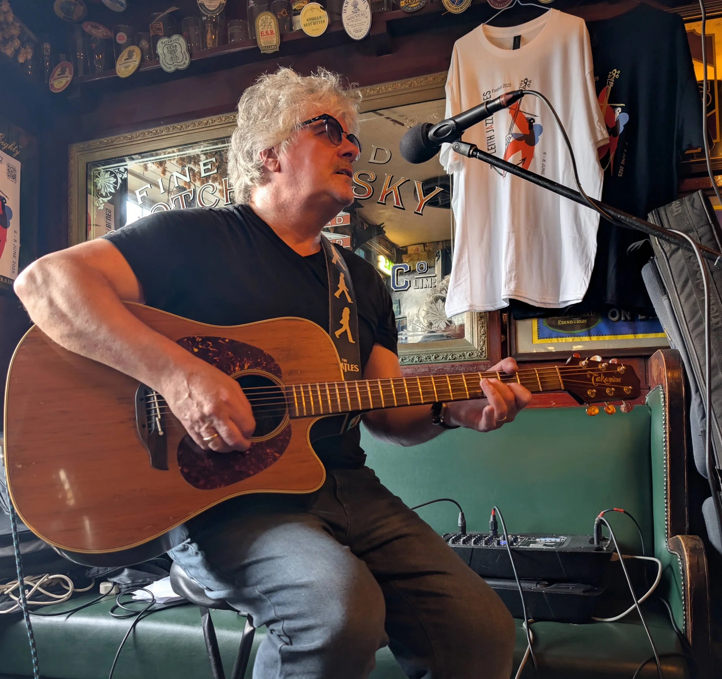 A man with curly gray hair and glasses playing an acoustic guitar and singing into a microphone in a bar. Behind him are T-shirts hanging on the wall and a mirror reflecting signs and decorations.