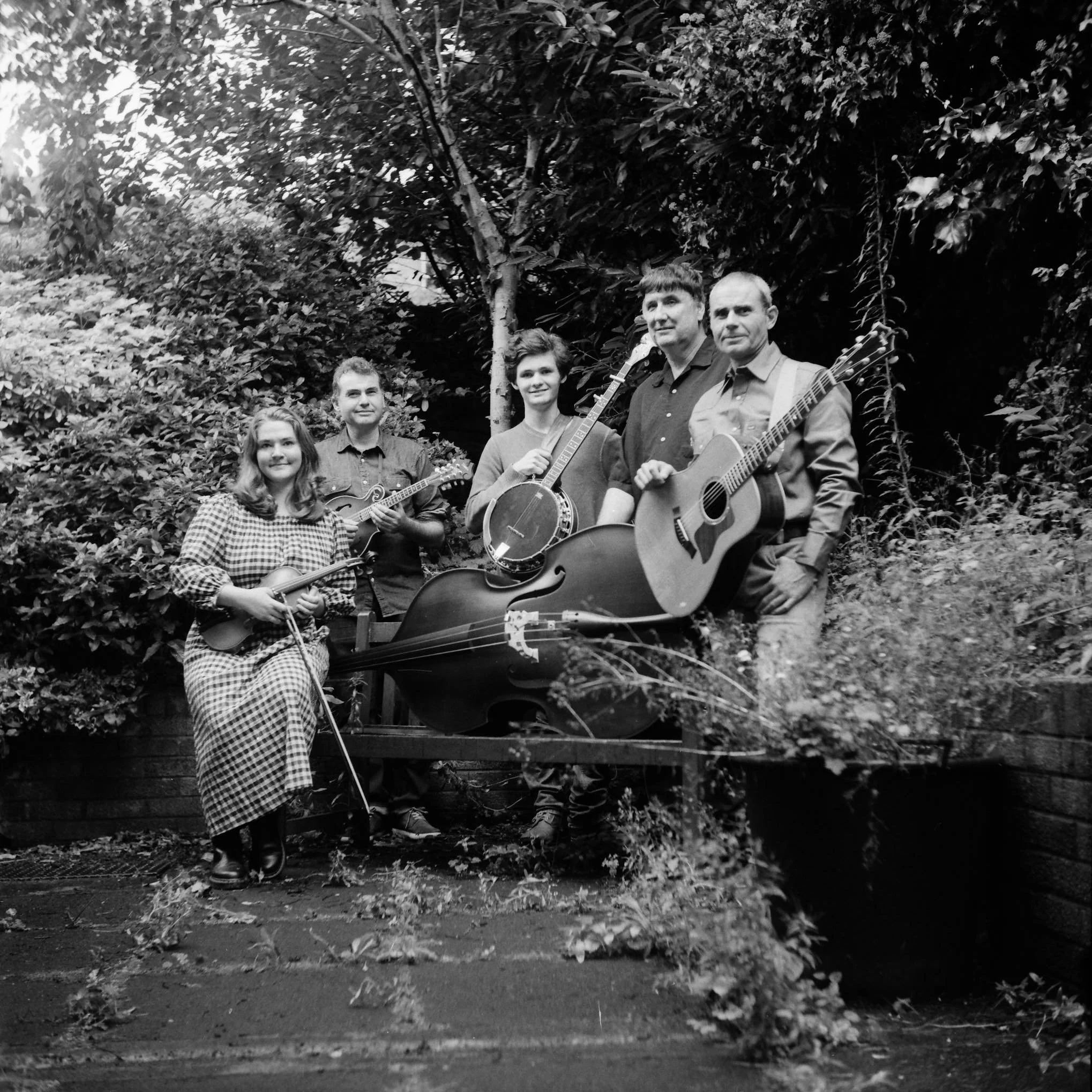 A group of six people outdoors amongst trees, with some holding musical instruments, including guitars and drums, and smiling at the camera.