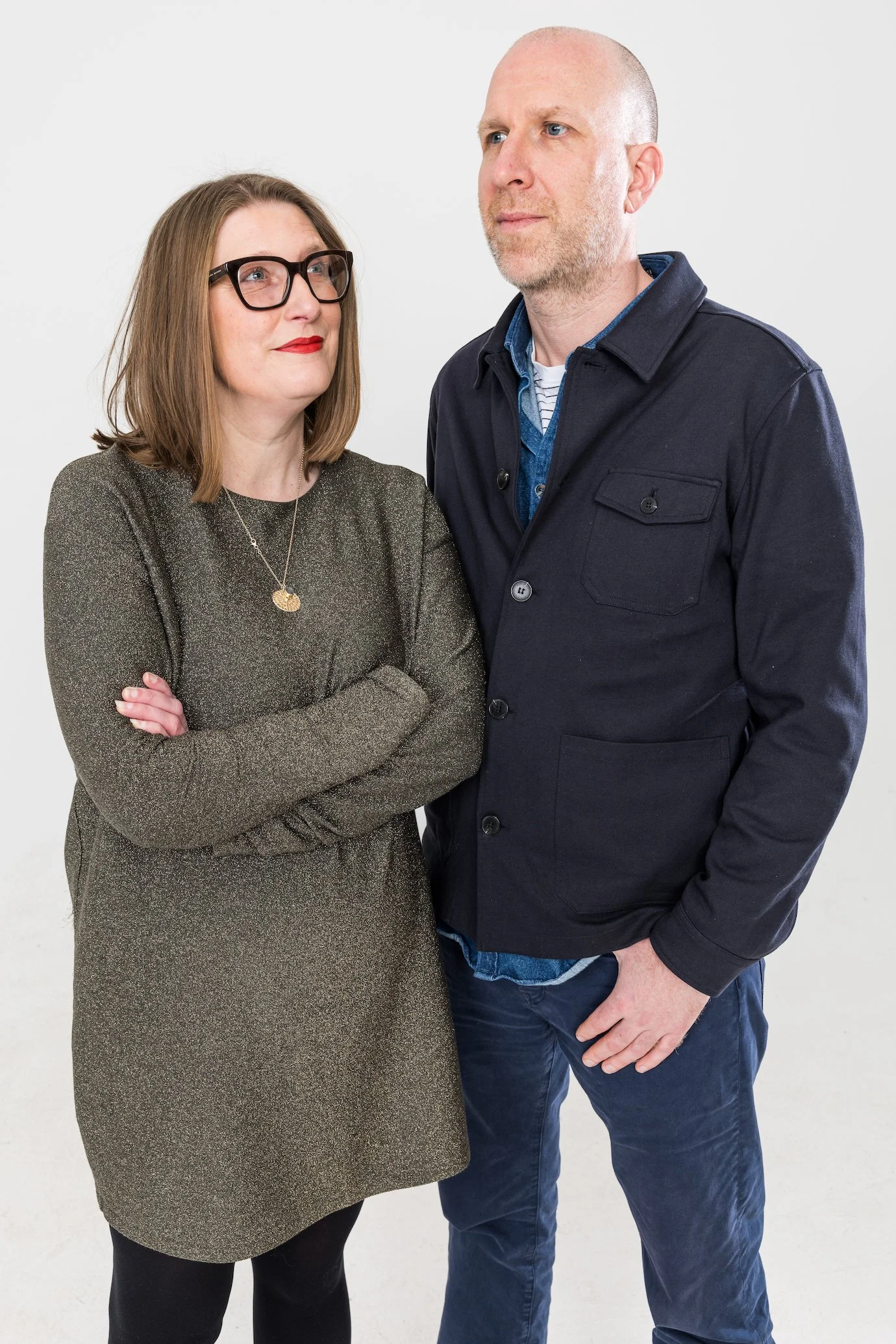 A woman with glasses and red lipstick standing with arms crossed, looking at a man with a beard and short hair, wearing a dark jacket, against a plain white background.