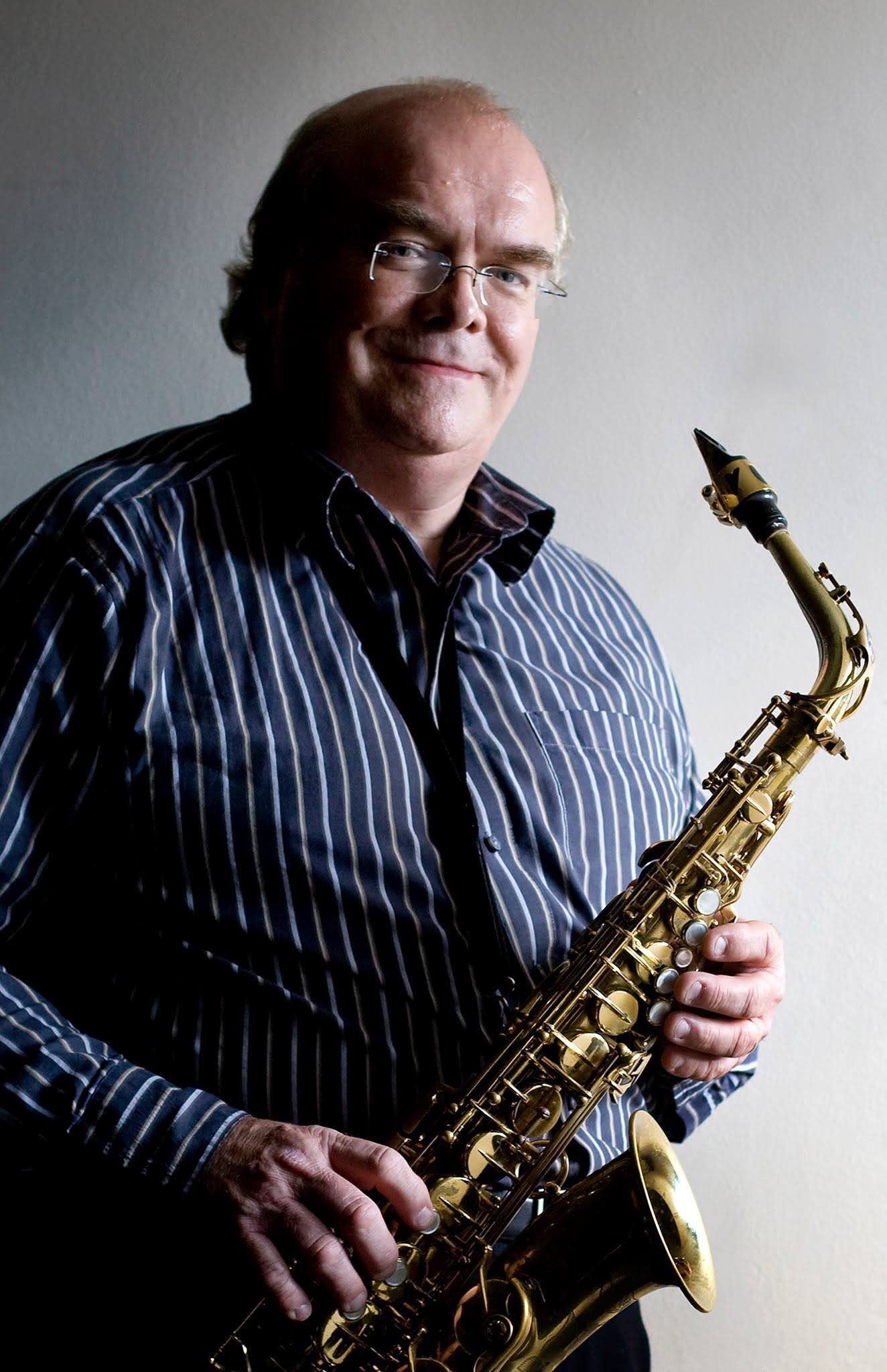 A man in a striped shirt holding a gold saxophone, standing in front of a plain wall.