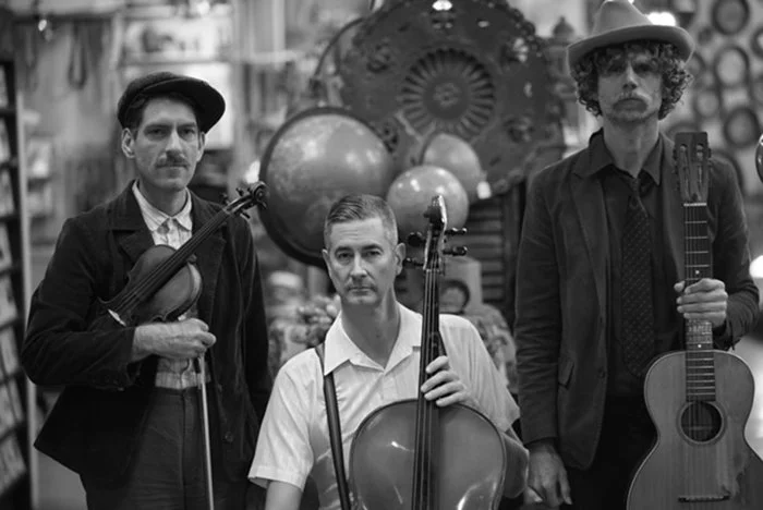 Three men standing in a store, each holding a musical instrument: a violin, a cello, and a guitar, with globe decorations in the background.