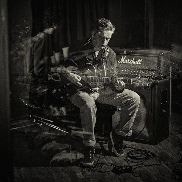 A man sitting on a stool playing an acoustic guitar in front of a Marshall amplifier in a music studio or rehearsal space.