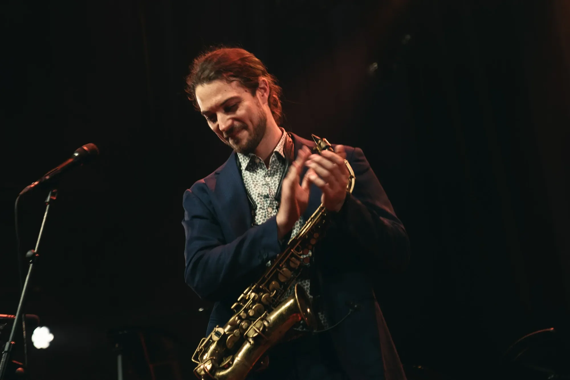 A man with long hair and a beard playing a saxophone on stage, dressed in a dark blazer and a patterned shirt, with a microphone in front of him and stage lights in the background.