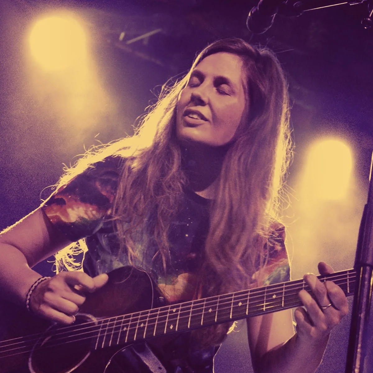 A woman with long wavy hair is playing an acoustic guitar on stage, illuminated by warm stage lights. She appears to be singing or immersed in music.