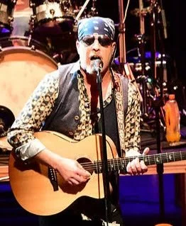 Musician performing on stage with guitar, wearing sunglasses, a bandana, and leopard print sleeves, with drums in the background.