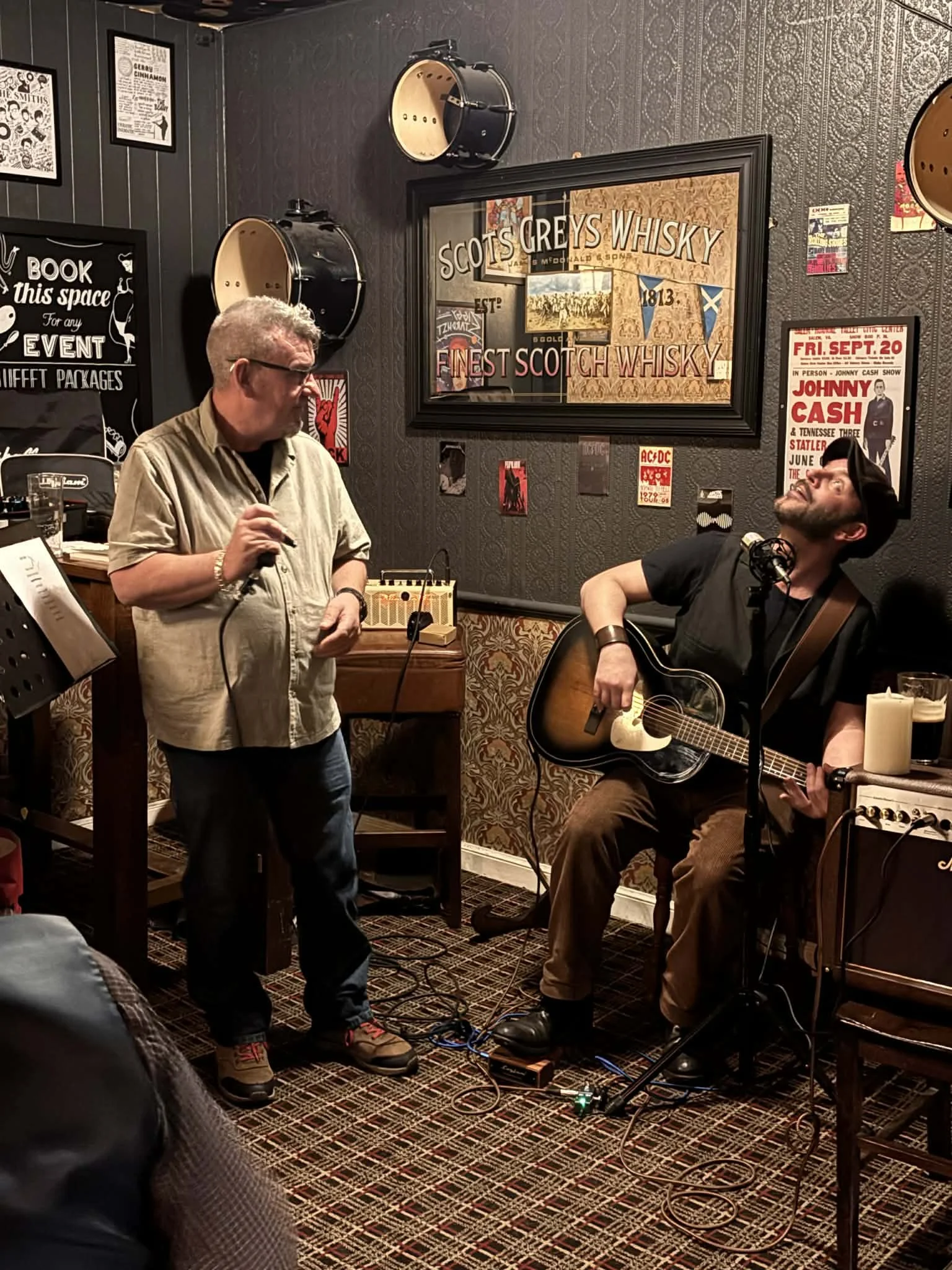 Two men performing traditional Scottish music in a pub or bar, with musical instruments including an acoustic guitar. One man is standing, and the other is sitting while playing the guitar. The background has vintage posters and signs, including one about Scottish whisky.