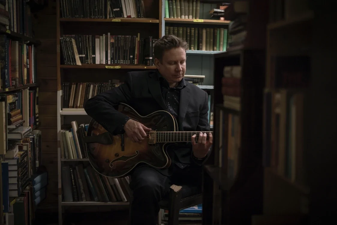 A man in a black suit playing an acoustic guitar in a library with shelves of books around him.