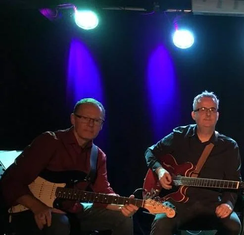 Two men playing electric guitars on stage with blue and purple stage lighting.
