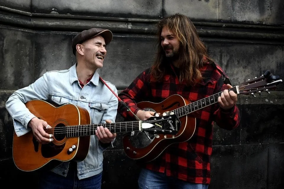 Two men playing acoustic guitars outdoors, smiling and enjoying their music.