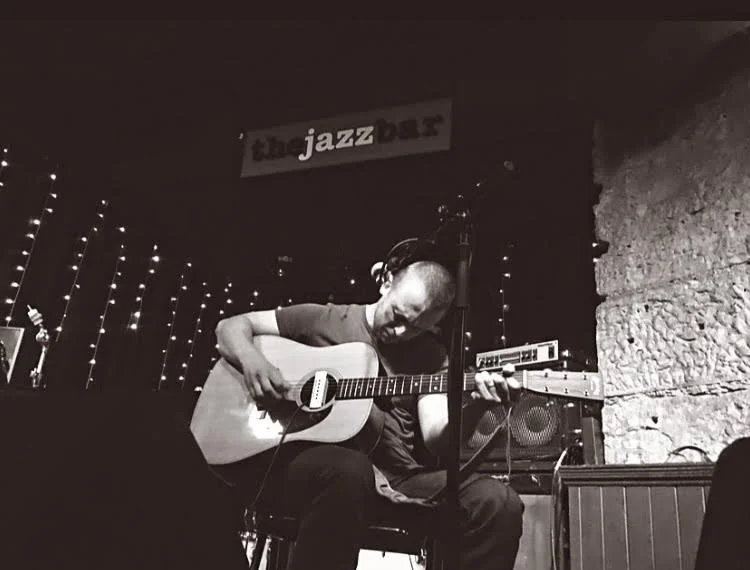 Man playing an acoustic guitar on stage at a jazz bar, with string lights and a brick wall in the background.