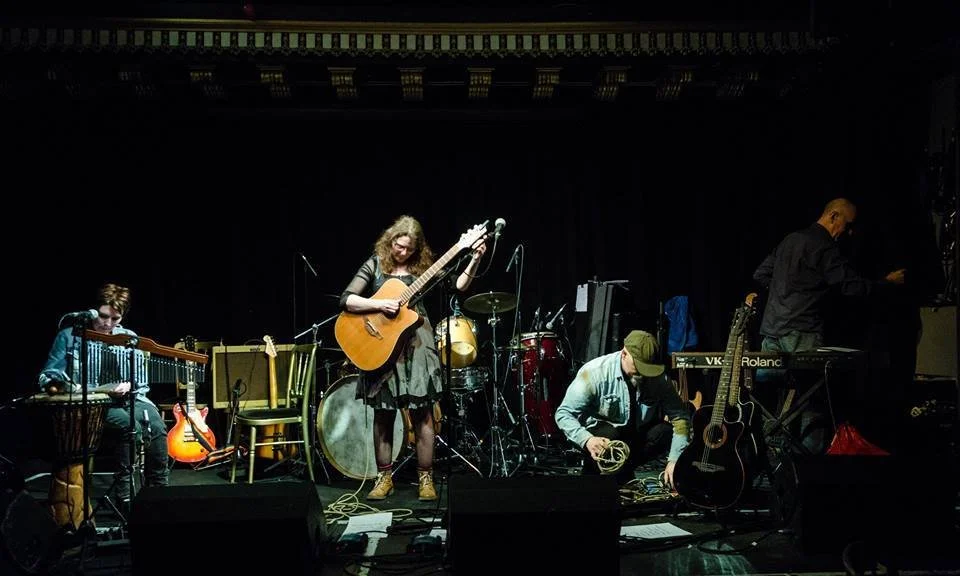 Musicians preparing for a performance in a dimly lit venue, with instruments including guitars, drums, keyboard, and percussion.