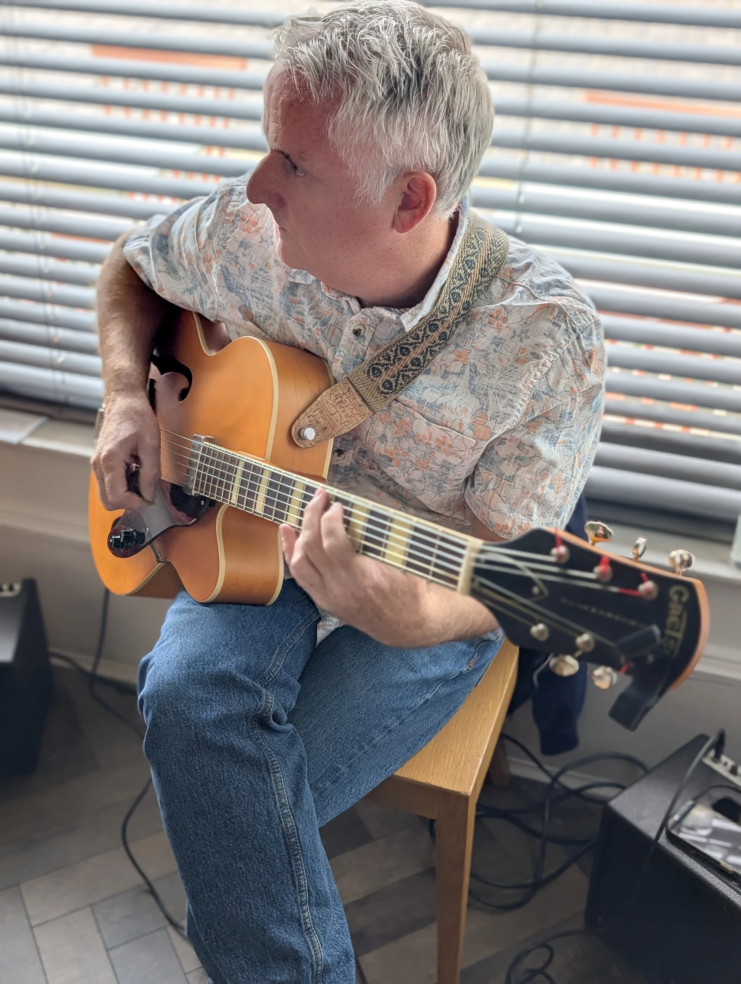 A man with gray hair sitting on a wooden stool, playing an acoustic guitar with a floral patterned shirt, in front of horizontal blinds.