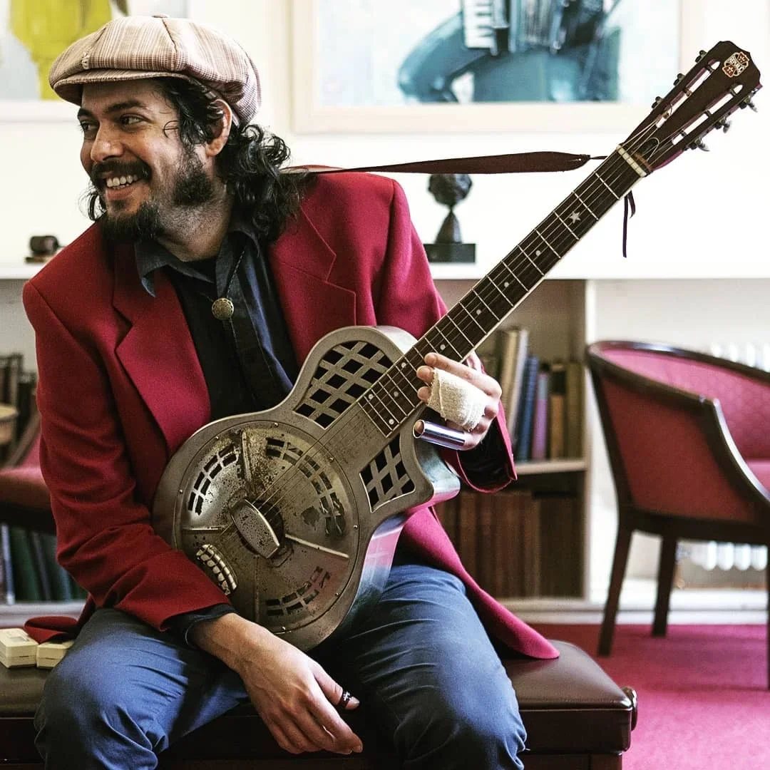 A man with long curly hair, a beard, and a hat, wearing a red blazer, playing a vintage metal resonator guitar in a cozy room with bookshelves and red chairs.