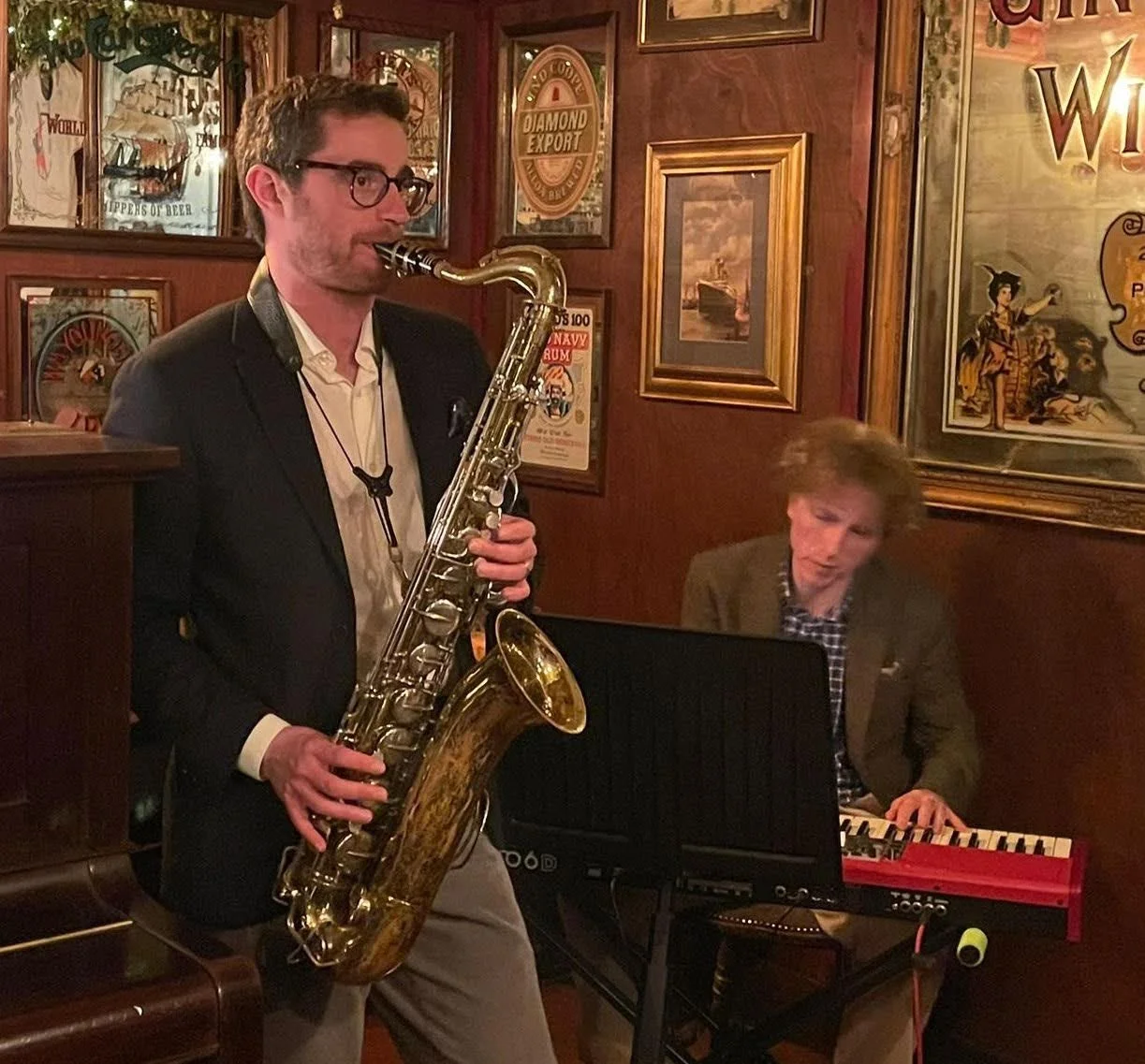 Two musicians performing in a cozy, vintage-style pub. The man in the foreground is playing a saxophone while wearing glasses and a suit jacket. The man in the background is playing a keyboard, dressed in a blazer and checked shirt. The pub's walls are decorated with framed vintage posters and artwork.