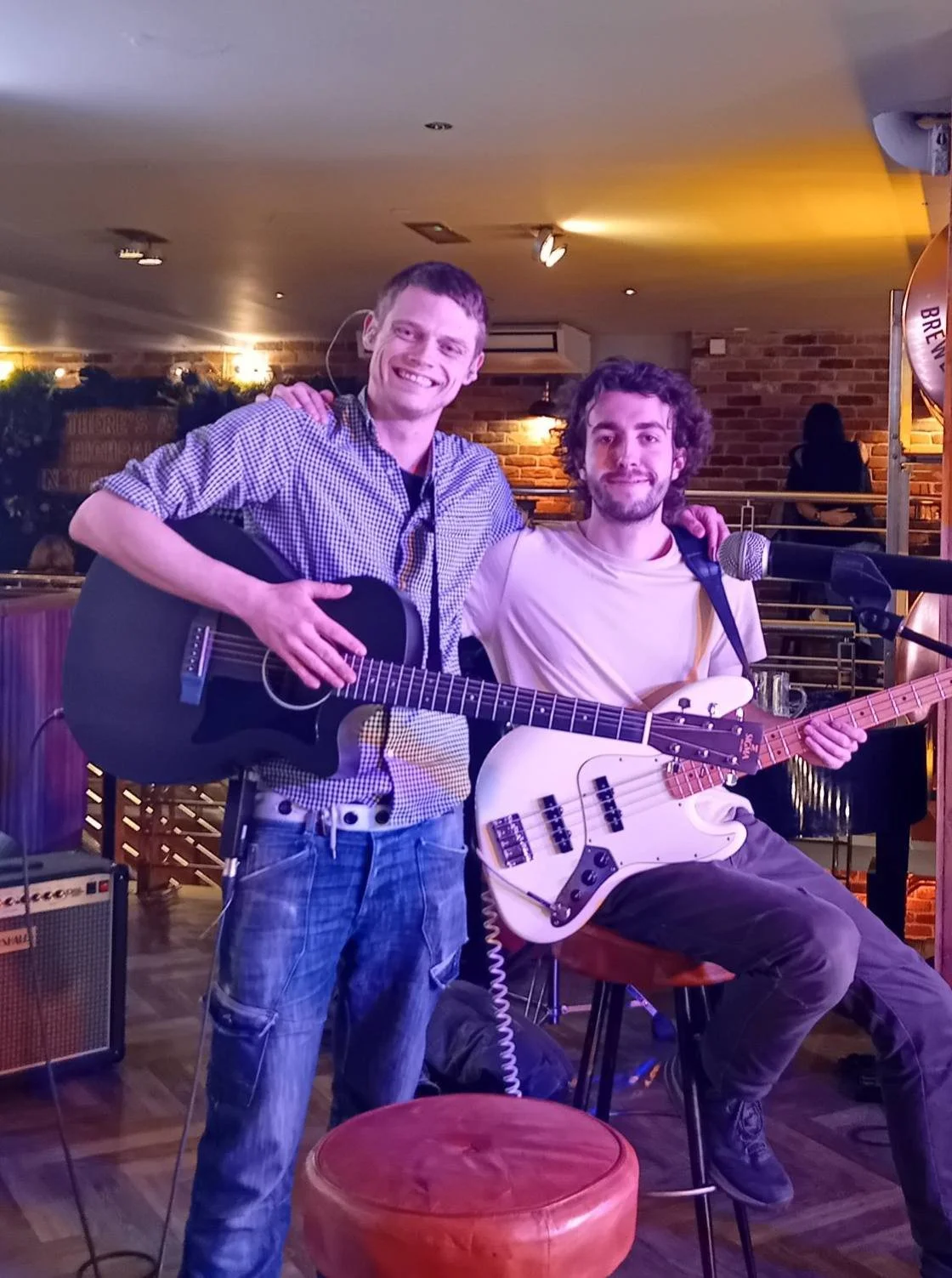 Two men with guitars posing for a photo in a cozy indoor setting, likely a bar or restaurant, with brick walls and warm lighting.