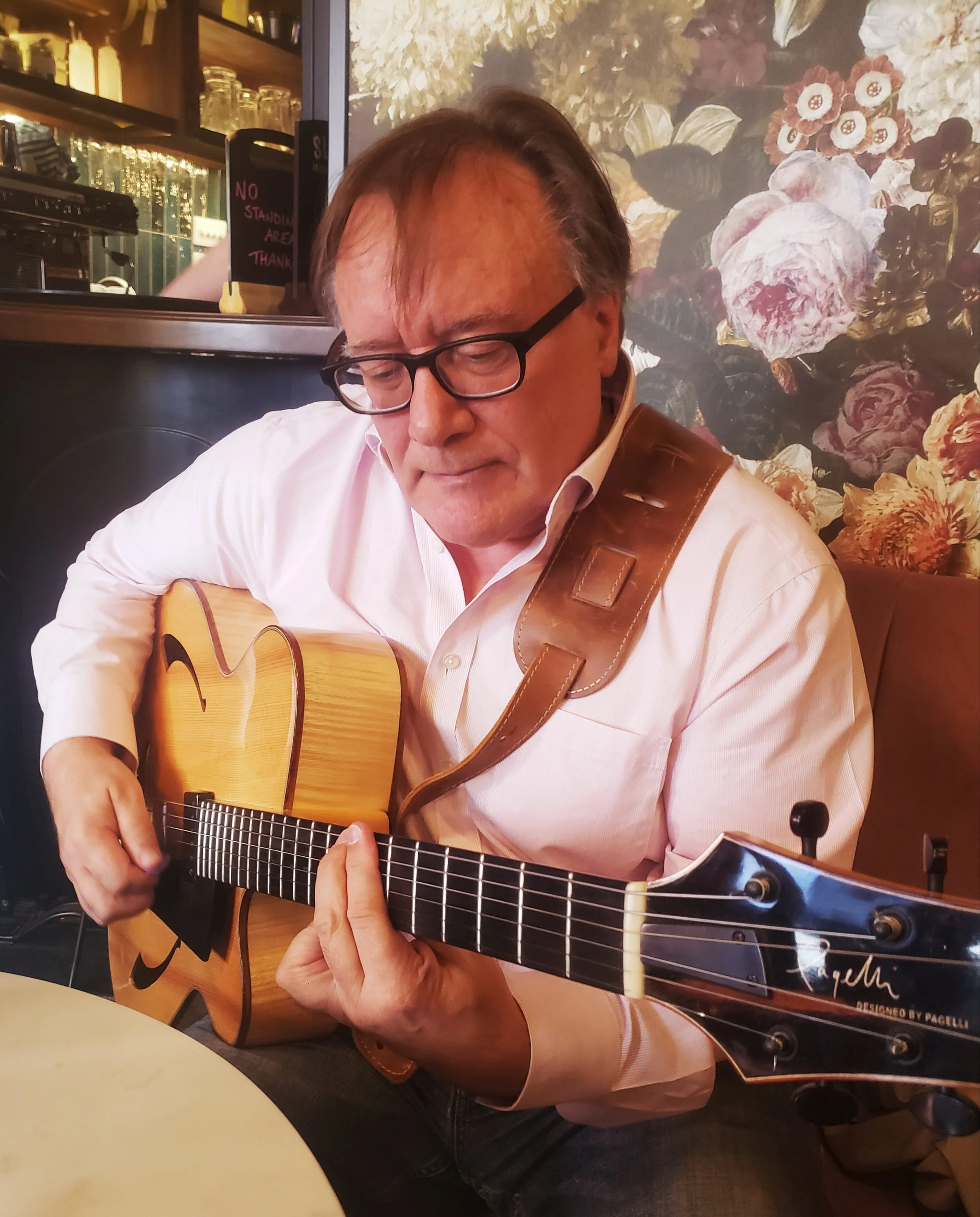 A man with glasses playing an acoustic guitar at a restaurant or cafe with floral wallpaper in the background.