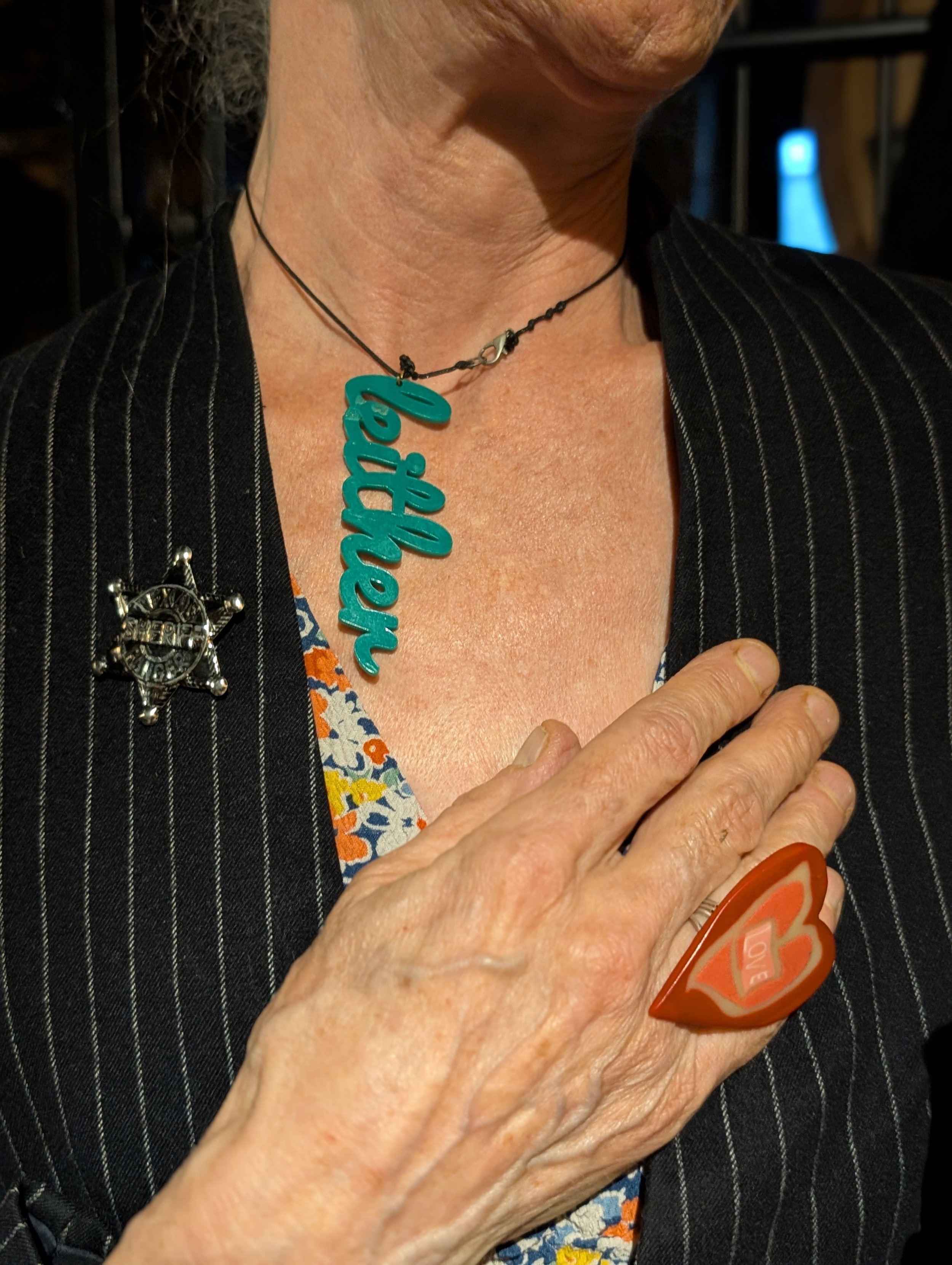Close-up of a person wearing a black striped blazer with sheriff badges, a colorful dress, a necklace with teal text, and holding a red heart-shaped object over their chest.