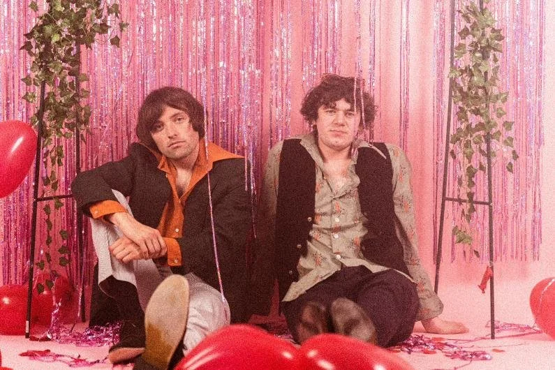 Two young men sitting on the floor in front of a pink, beaded curtain backdrop with heart-shaped balloons and pink decorations, suggesting a romantic or celebratory setting.