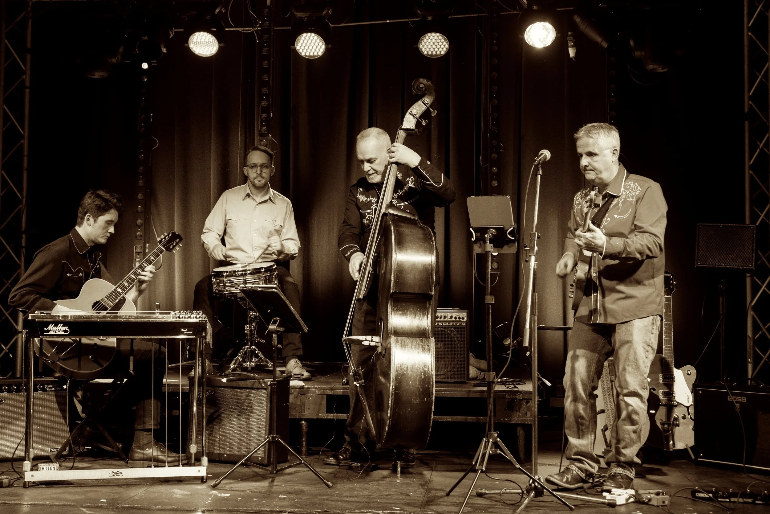 A four-piece band performing on stage with guitars, drums, and double bass, with stage lights overhead and a dark curtain background.
