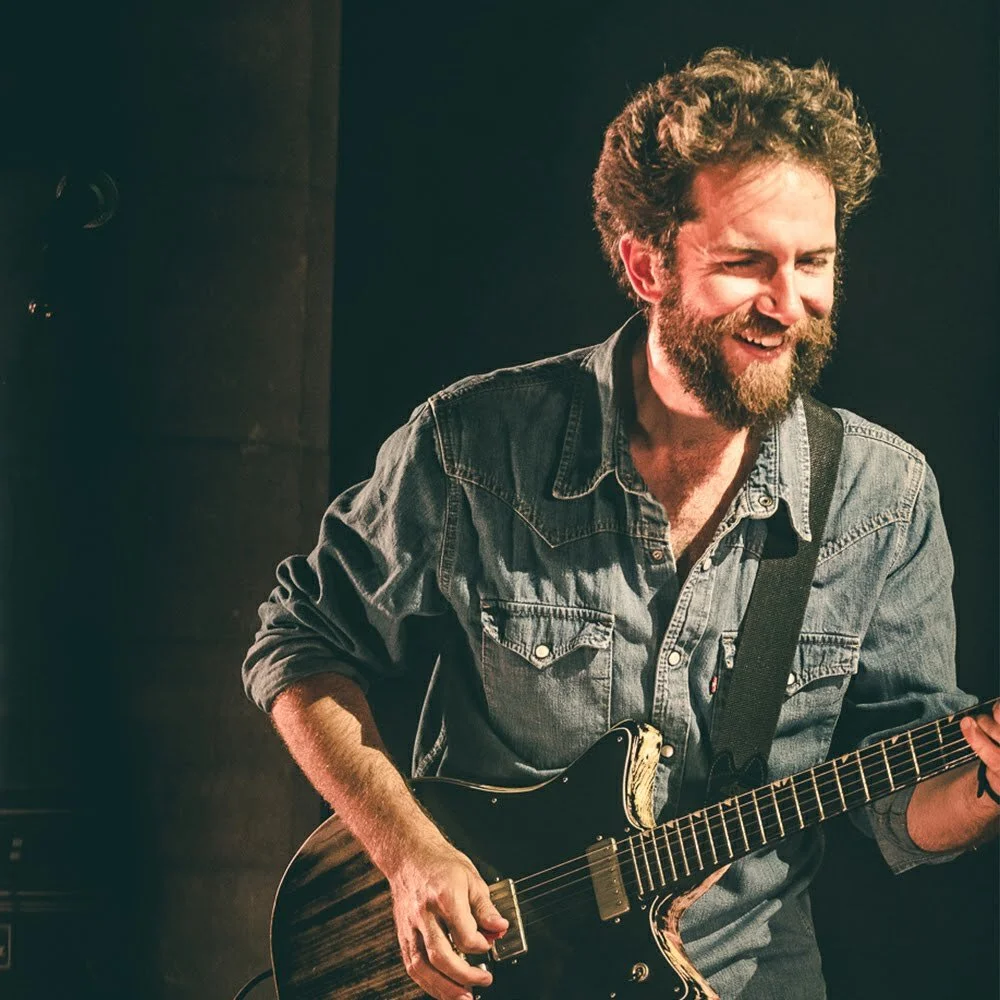 A man with curly hair and a beard playing an electric guitar and smiling.