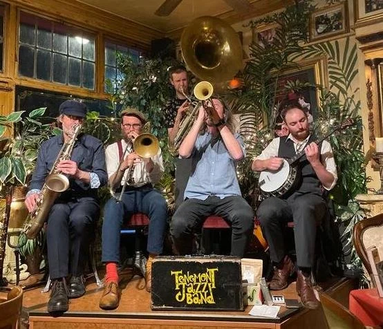 A jazz band performing indoors with four musicians playing saxophone, trombone, tuba, and banjo, seated in front of a yellow sign that reads "TéNEMONT jazz BAND" surrounded by plants and decorated walls.
