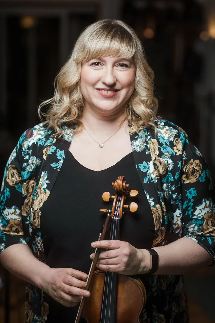 A woman with blonde curly hair holding a violin in her hands, smiling, wearing a black dress and a floral jacket.