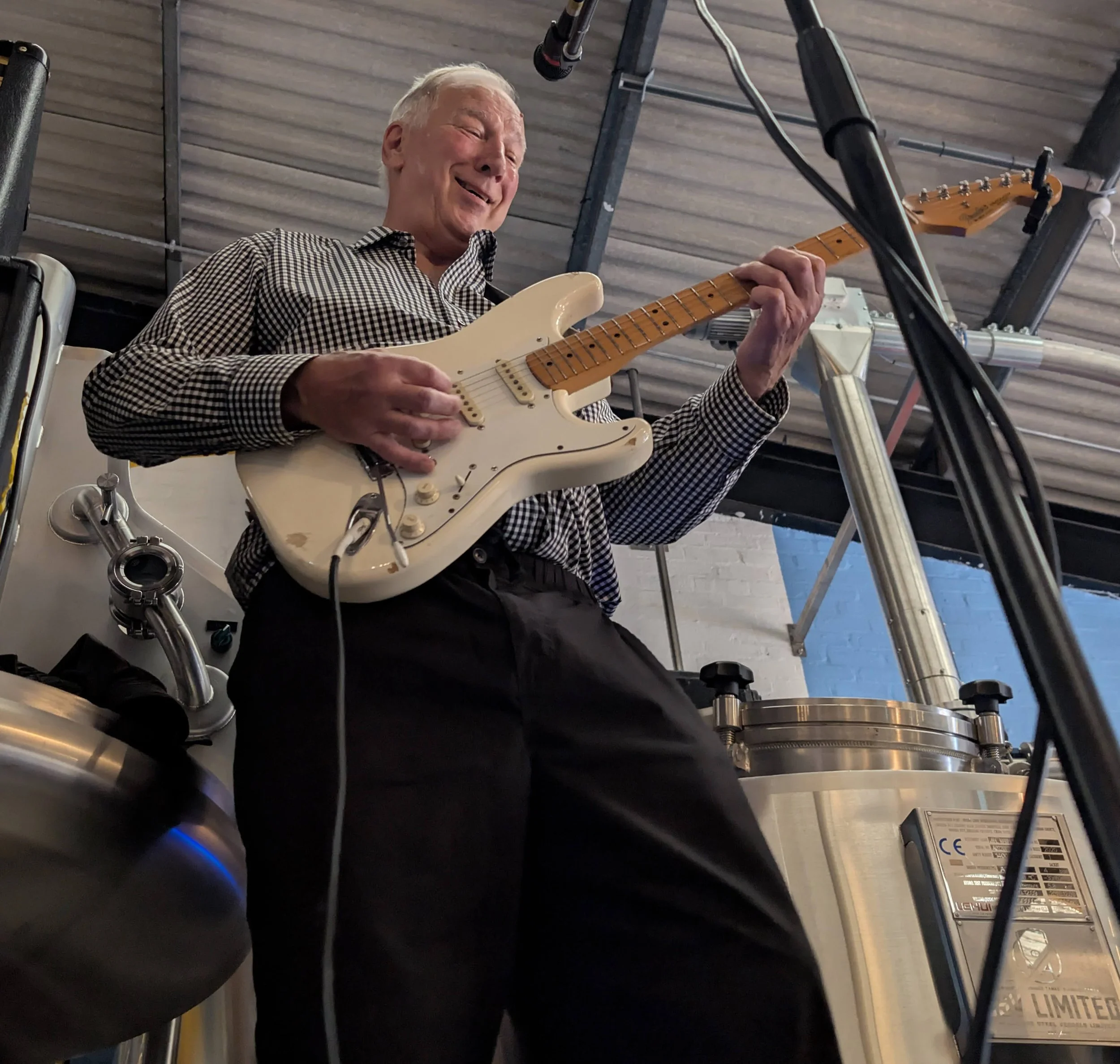 Elderly man playing an electric guitar in a brewery or industrial setting, smiling and enjoying the music.
