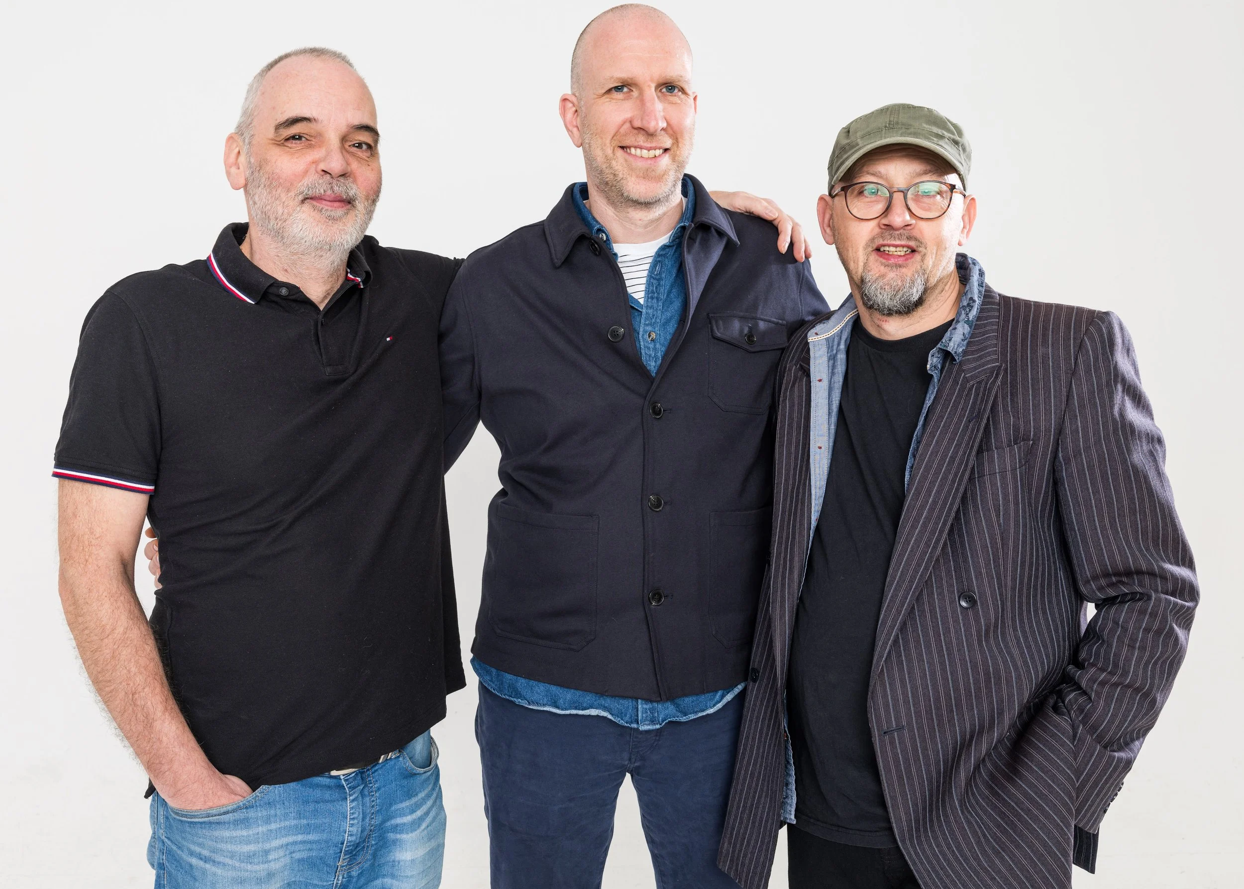 Three men standing close together, smiling, with one man in the middle having his arms around the others. All are casually dressed and looking at the camera against a plain white background.