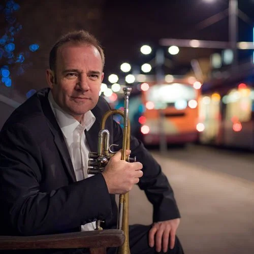 A man in a dark suit holding a trumpet, sitting outdoors at night with a blurred city bus and lights in the background.