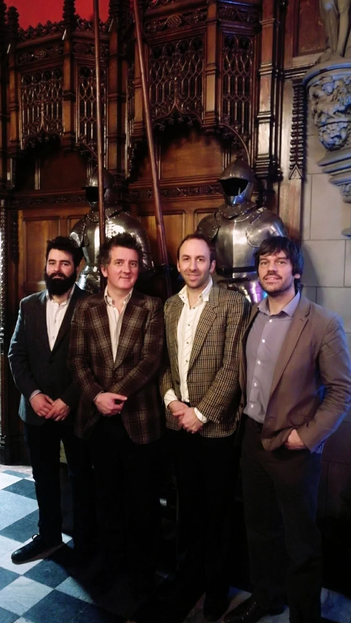 Four men in suits standing in front of ornate wooden and stone wall with suits of armor behind them.