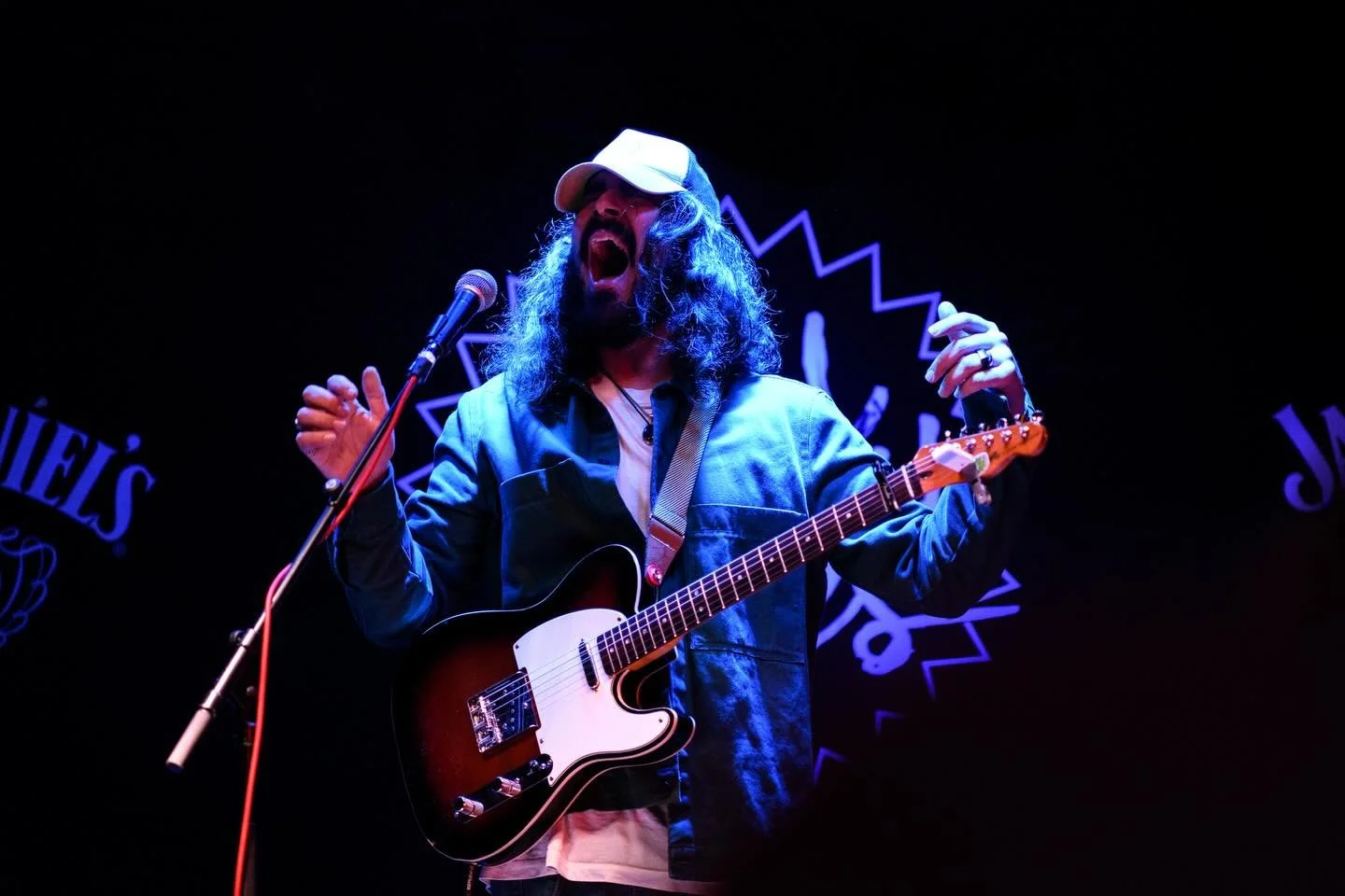 Man with long curly hair, beard, and glasses singing into a microphone, playing an electric guitar on stage with blue and purple lighting.