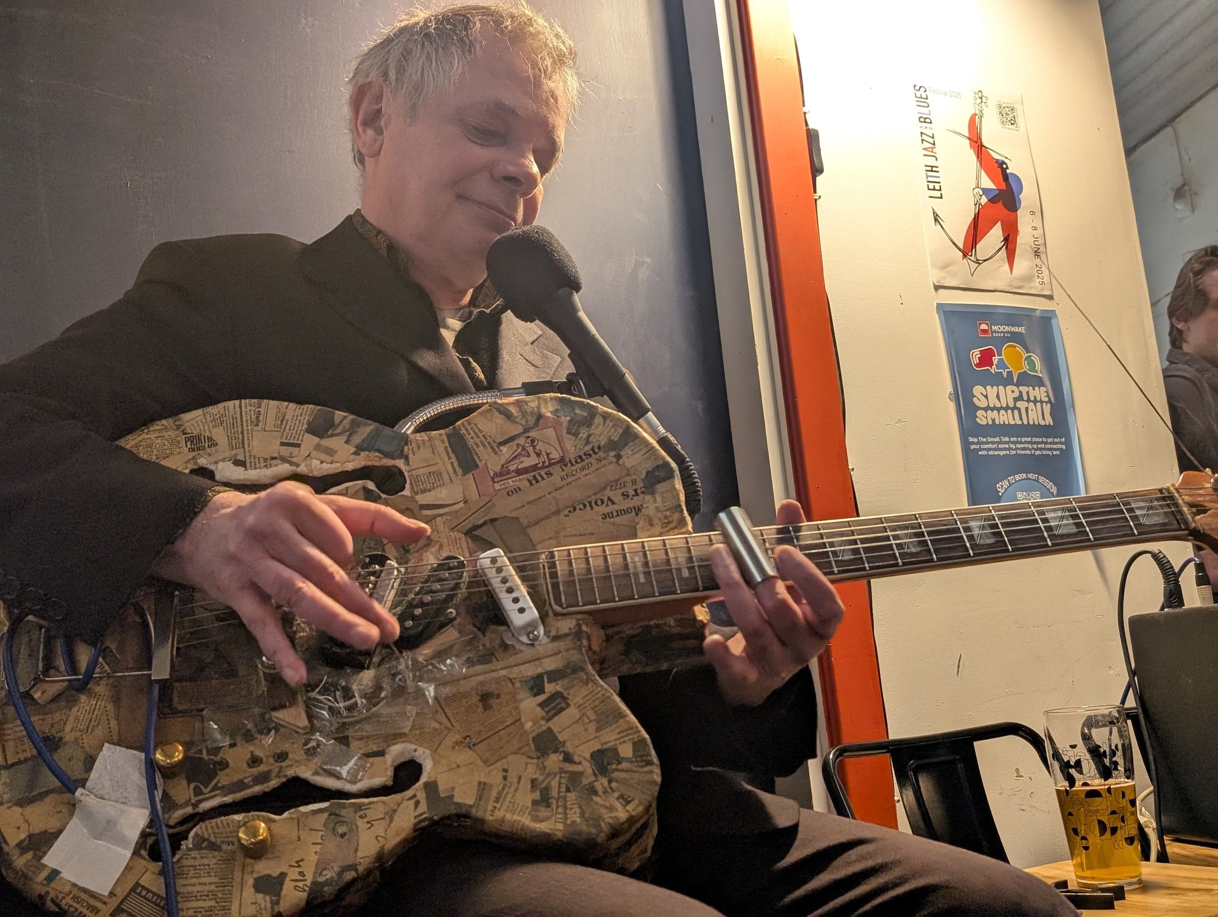 A man playing an guitar, sitting in front of a microphone, with posters on the wall behind him.