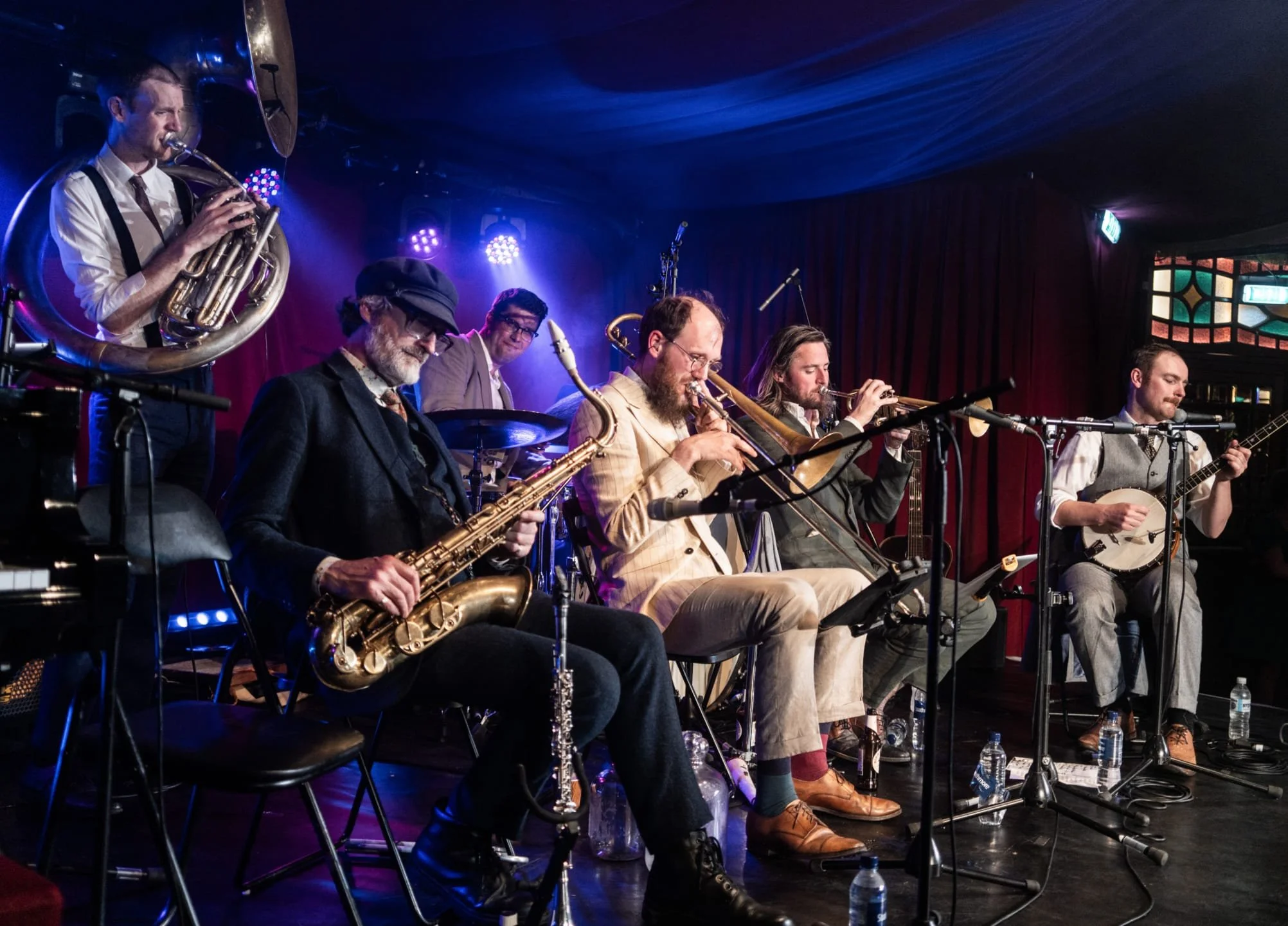 A jazz band performing on stage with musicians playing saxophones, trumpet, and trombone, while a singer plays the guitar. The stage has dark curtains, colorful lighting, and a stained-glass window in the background.